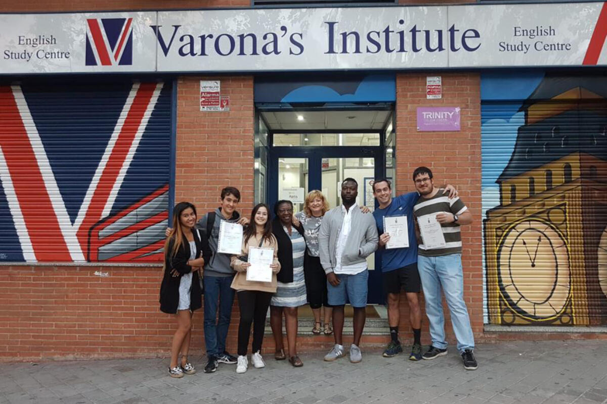 Un grupo de ocho personas sonrientes posa frente al Instituto Varona, con sus certificados en la mano. El edificio luce un mural con la bandera británica. El ambiente es festivo.