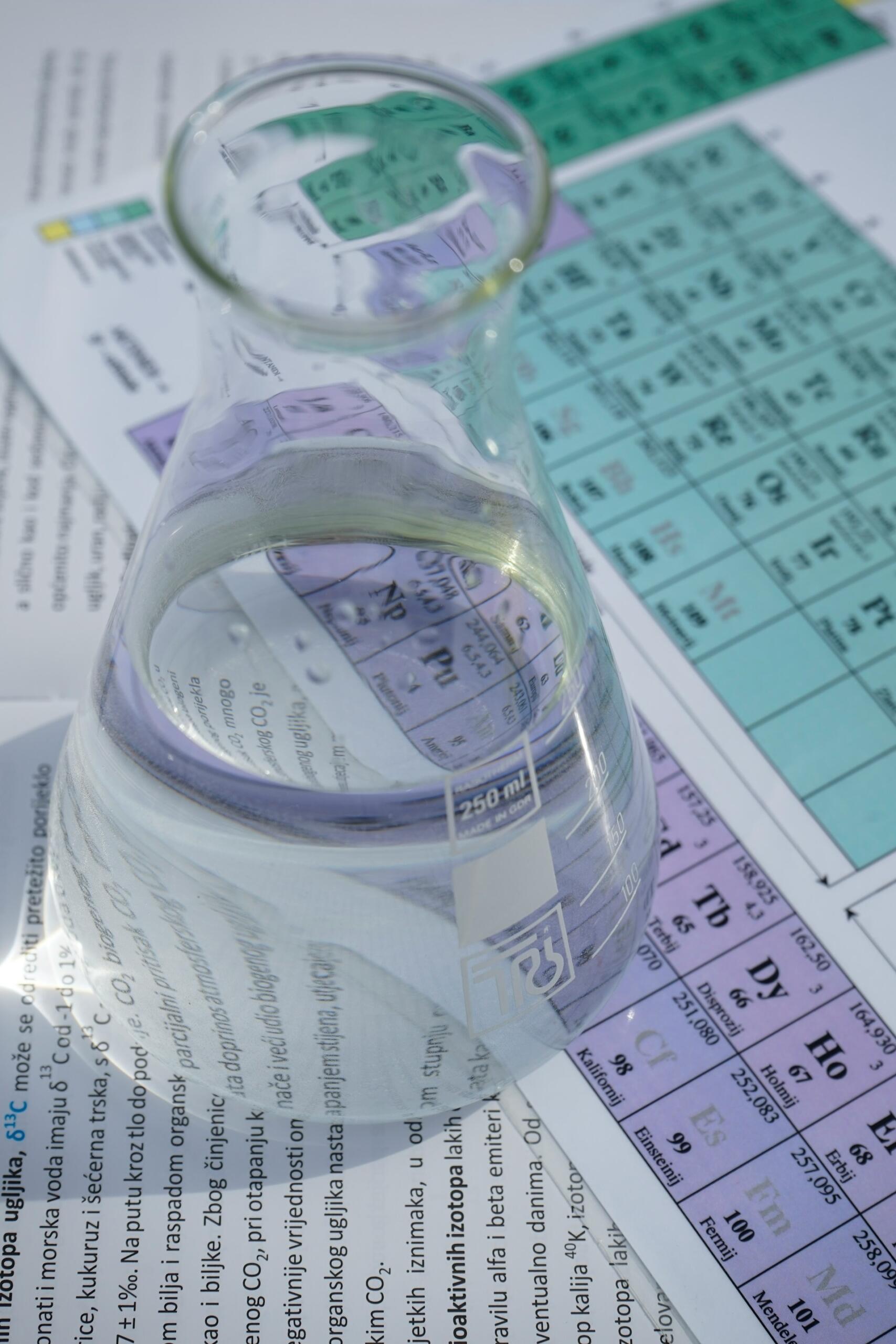 Un matraz Erlenmeyer de vidrio lleno de líquido transparente se encuentra sobre una tabla periódica de elementos, creando una atmósfera científica y educativa.