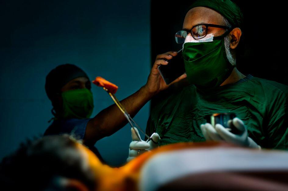 Un cirujano con bata verde y mascarilla habla por teléfono durante una operación, sosteniendo un instrumento. El ambiente es tenso, con una concentración absoluta.