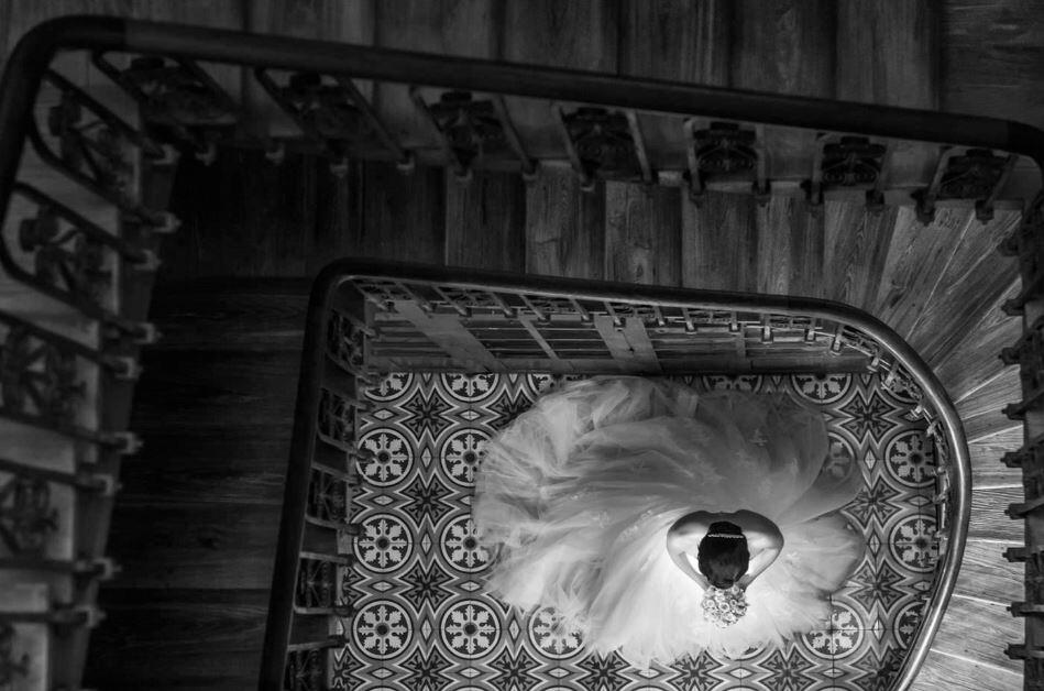 Imagen en blanco y negro de una novia vista desde arriba, de pie sobre un suelo de baldosas ornamentadas dentro de una escalera de caracol. Su vestido se extiende en forma circular.