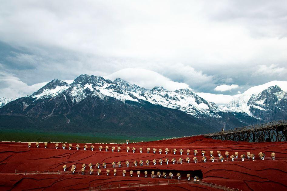 Fila de artistas con trajes tradicionales en un escenario rojo con gradas, con montañas nevadas al fondo, bajo un cielo nublado. Majestuoso y dramático.