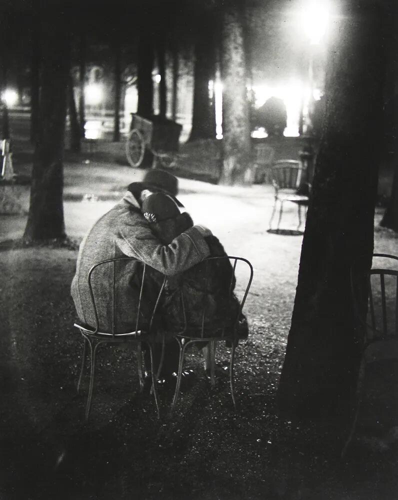Una pareja se sienta en unas sillas bajo los árboles en un parque tenuemente iluminado, abrazándose con ternura. La escena es serena e íntima, y transmite una sensación de tranquilo romanticismo.