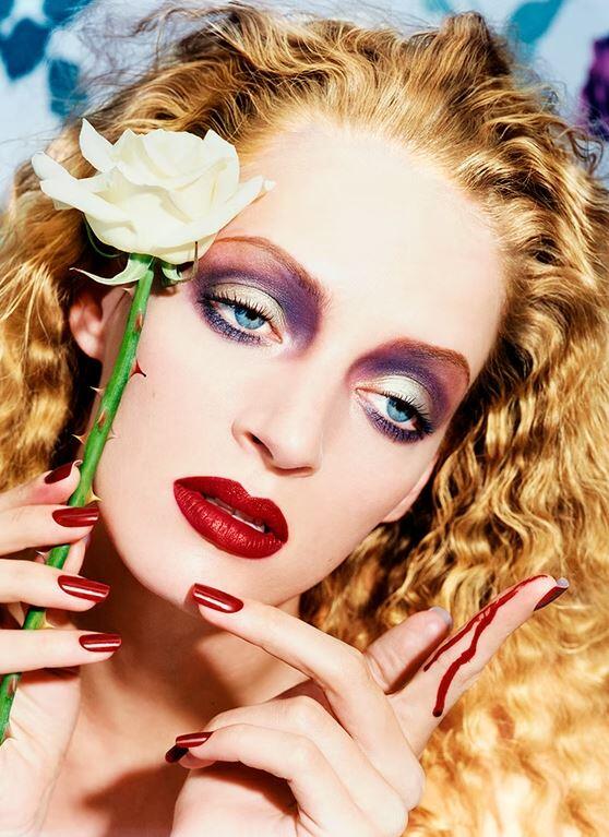 Uma Thurman con un maquillaje dramático sostiene una rosa blanca. Su expresivo rostro muestra unos labios rojos llamativos y una sombra de ojos intensa. Tiene el pelo largo y rizado y un corte en el dedo.