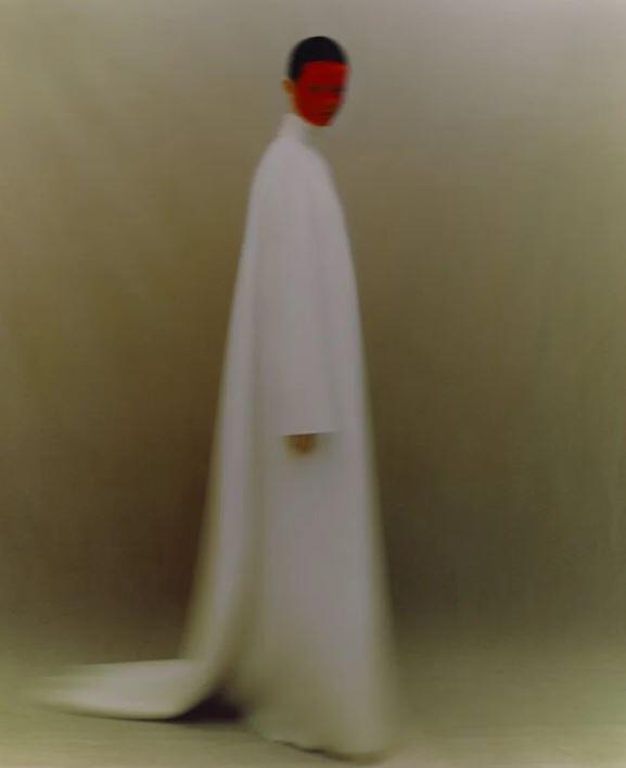 Una persona con una túnica blanca larga y fluida se encuentra de pie frente a un fondo difuminado y neutro. La cara está pintada de rojo, lo que crea una atmósfera misteriosa y surrealista.