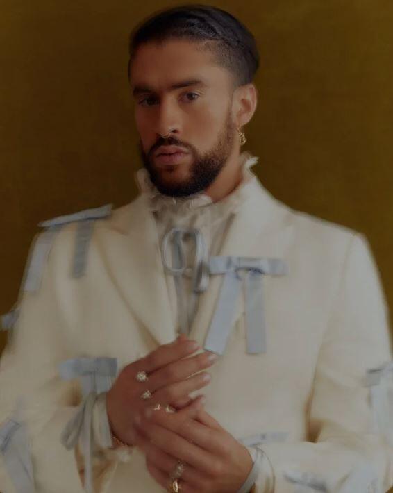 Bad Bunny vestido con un traje blanco adornado con cintas azul claro posa con confianza. Lleva barba, el pelo peinado, pendientes y varios anillos, sobre un fondo color mostaza.