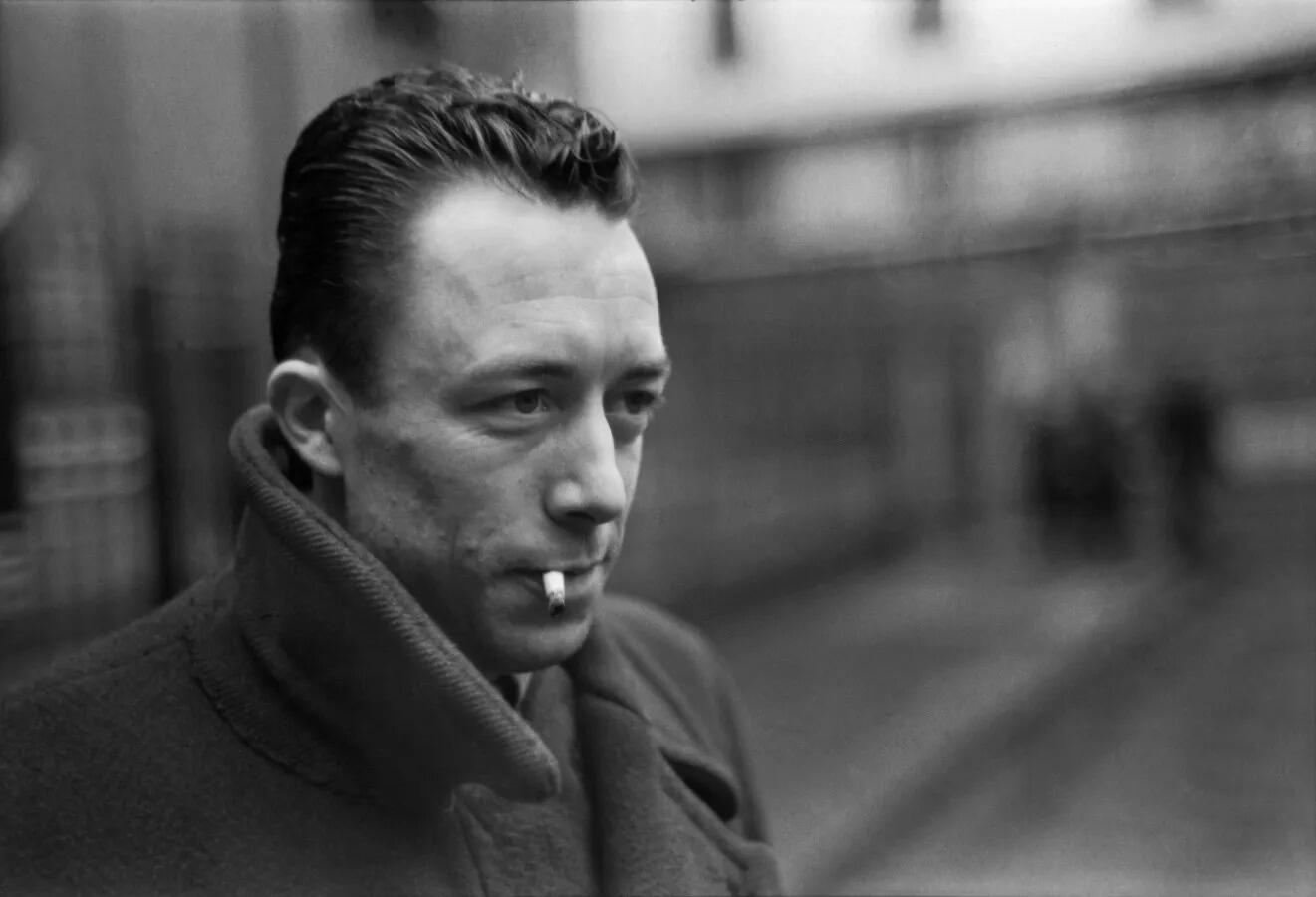 Fotografía en blanco y negro de Albert Camus con un cigarrillo en la boca, vestido con un abrigo grueso. Mira pensativo, con un fondo urbano borroso.
