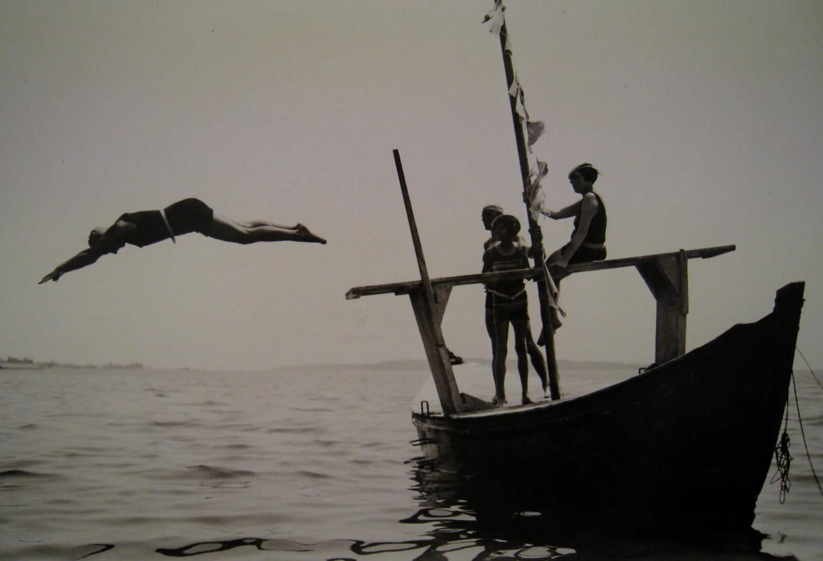 Una imagen en tonos sepia muestra a una persona saltando al mar desde una pequeña embarcación. Tres personas en la embarcación observan, rodeadas por aguas tranquilas y un cielo sereno.