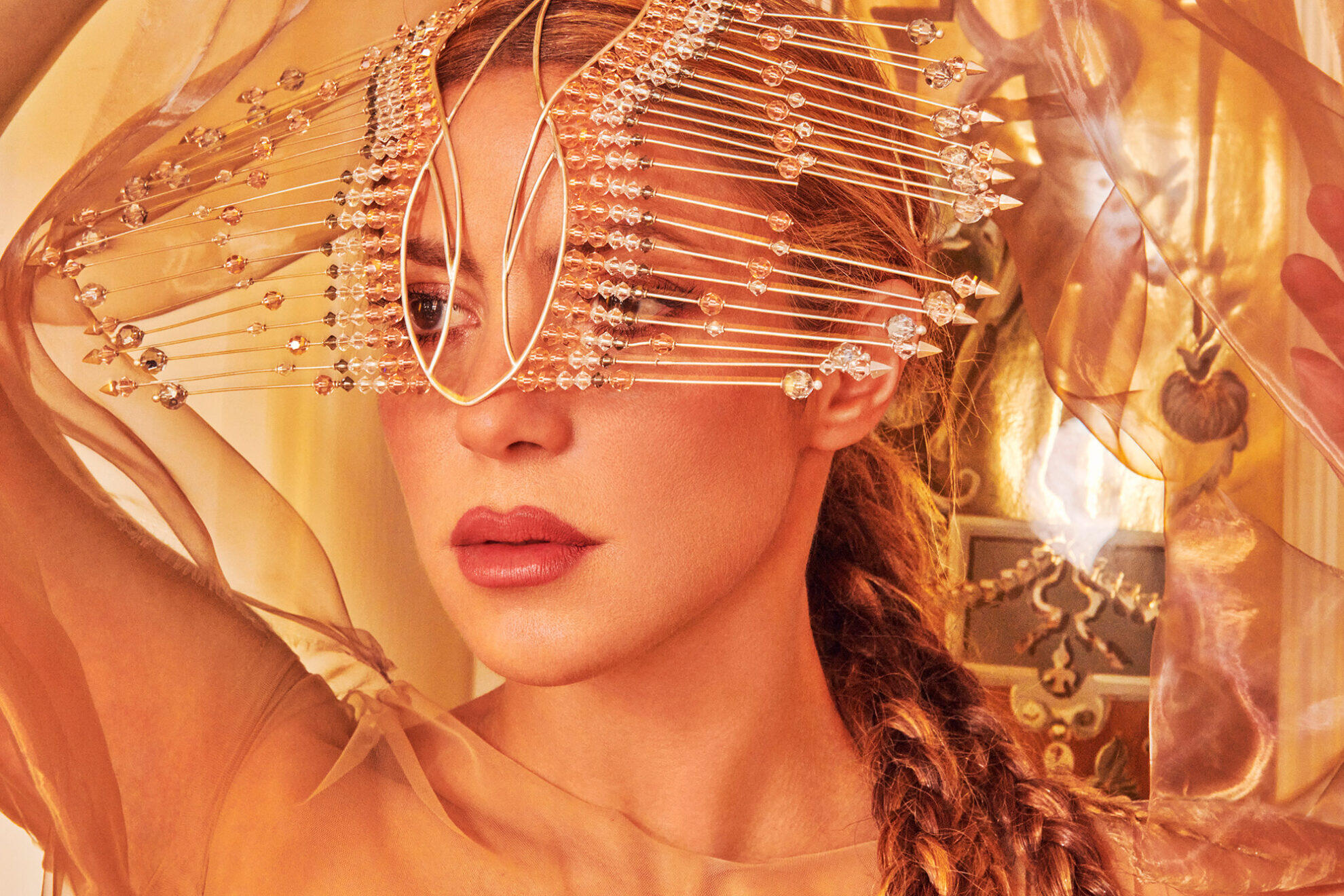 Shakira con expresión serena lleva un intrincado tocado de finos alambres metálicos y cristales, sobre un fondo dorado y etéreo.
