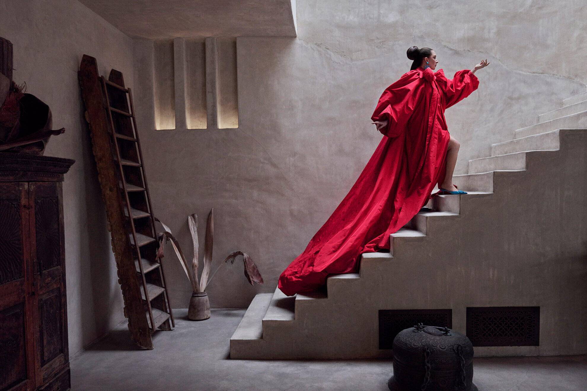 Maya Rudolph con un elegante vestido rojo con una larga cola sube con elegancia una escalera minimalista. La habitación está tenuemente iluminada con tonos tierra, lo que le confiere un ambiente sereno y dramático.