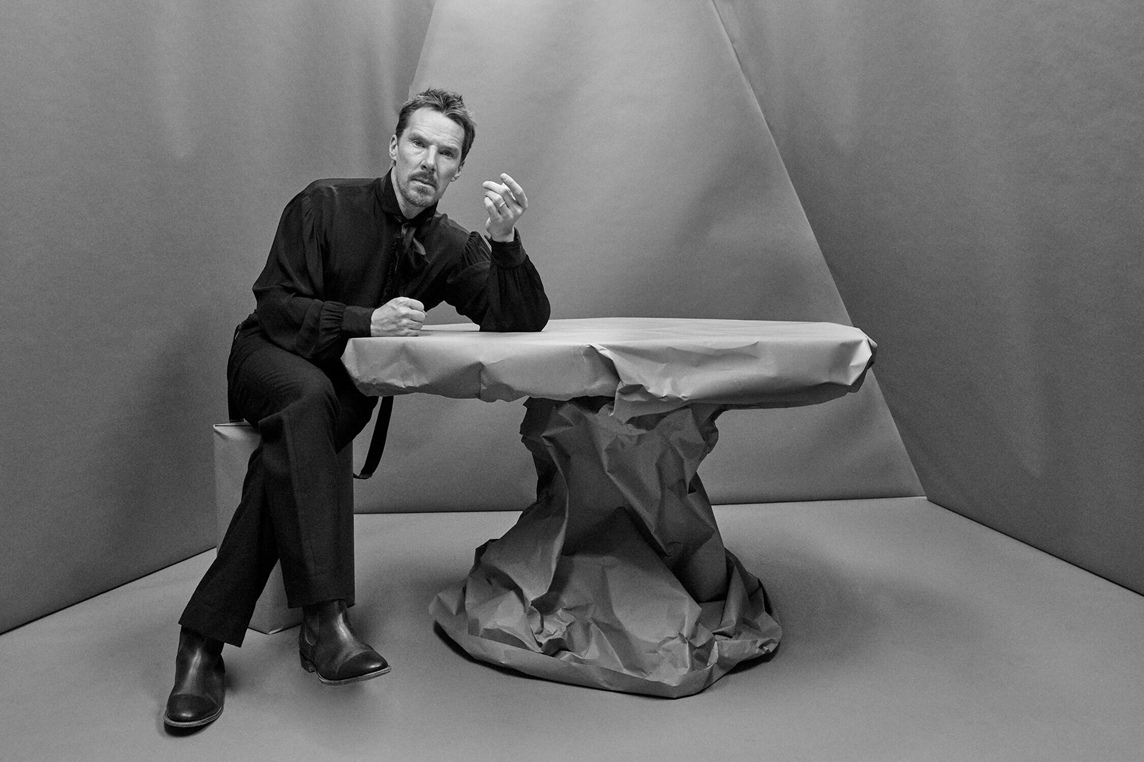 Imagen en blanco y negro de Benedict Cumberbatch vestido con ropa oscura sentado junto a una mesa cubierta de papeles arrugados. El fondo geométrico le da un toque moderno y artístico.