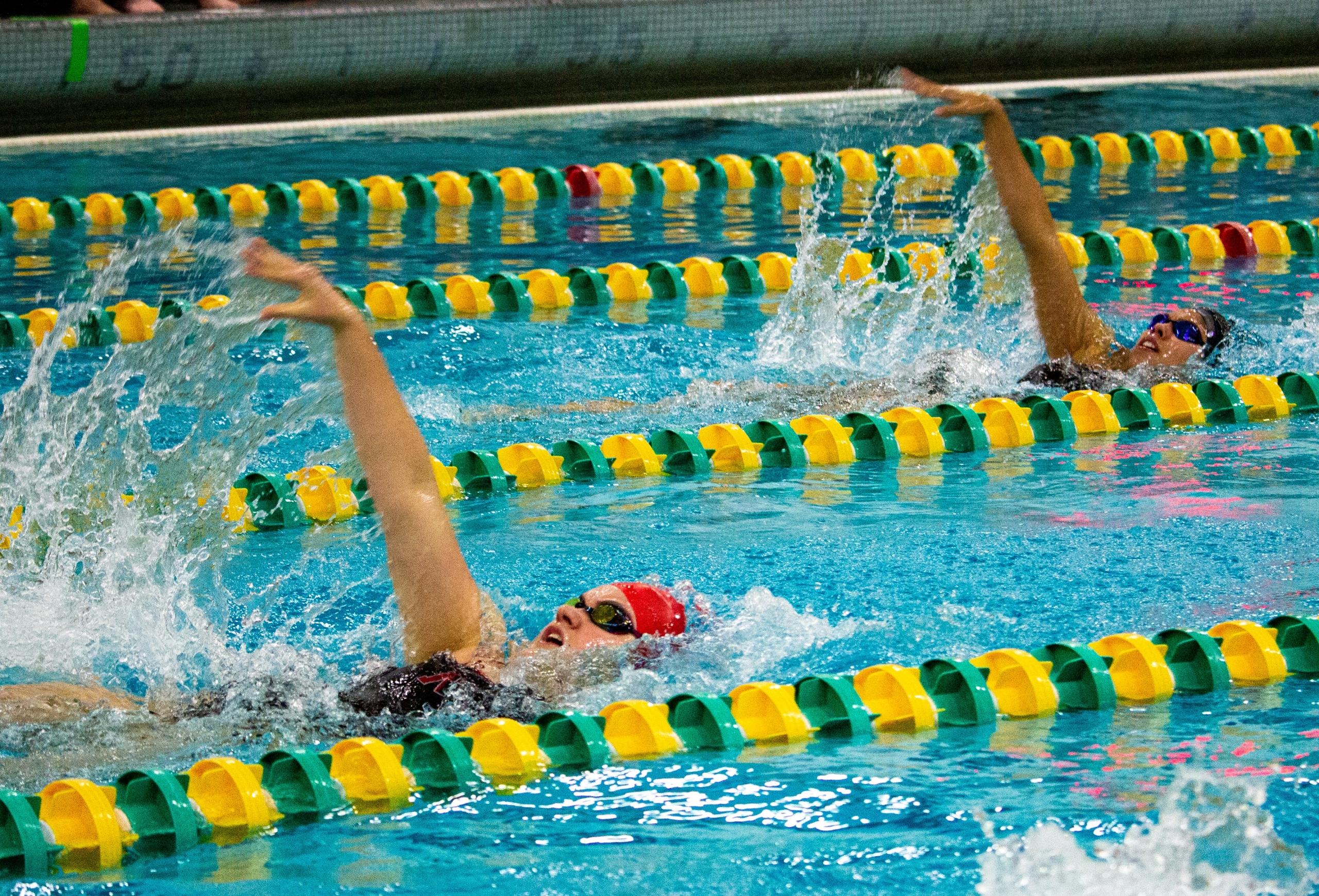 Dos nadadores competitivos nadan de espaldas en una piscina, creando salpicaduras, con marcadores de calle brillantes en primer plano.
