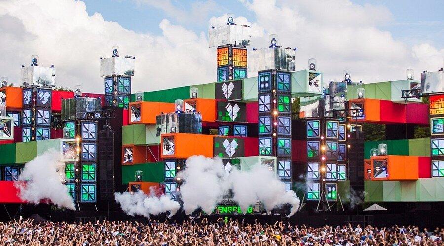Un enorme escenario de festival con coloridas pantallas geométricas y luces vibrantes, rodeado por una gran multitud bajo un cielo parcialmente nublado, que emite vibraciones energéticas.