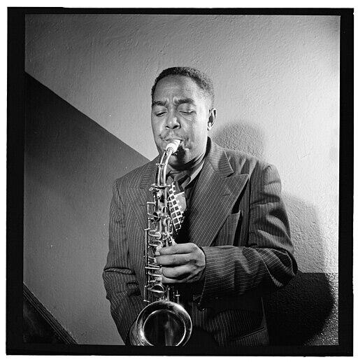Un Charlie Parker toca el saxofón con los ojos cerrados, vestido con un traje a rayas. El escenario tiene un fondo minimalista y sombreado, que transmite una profunda concentración y emoción.