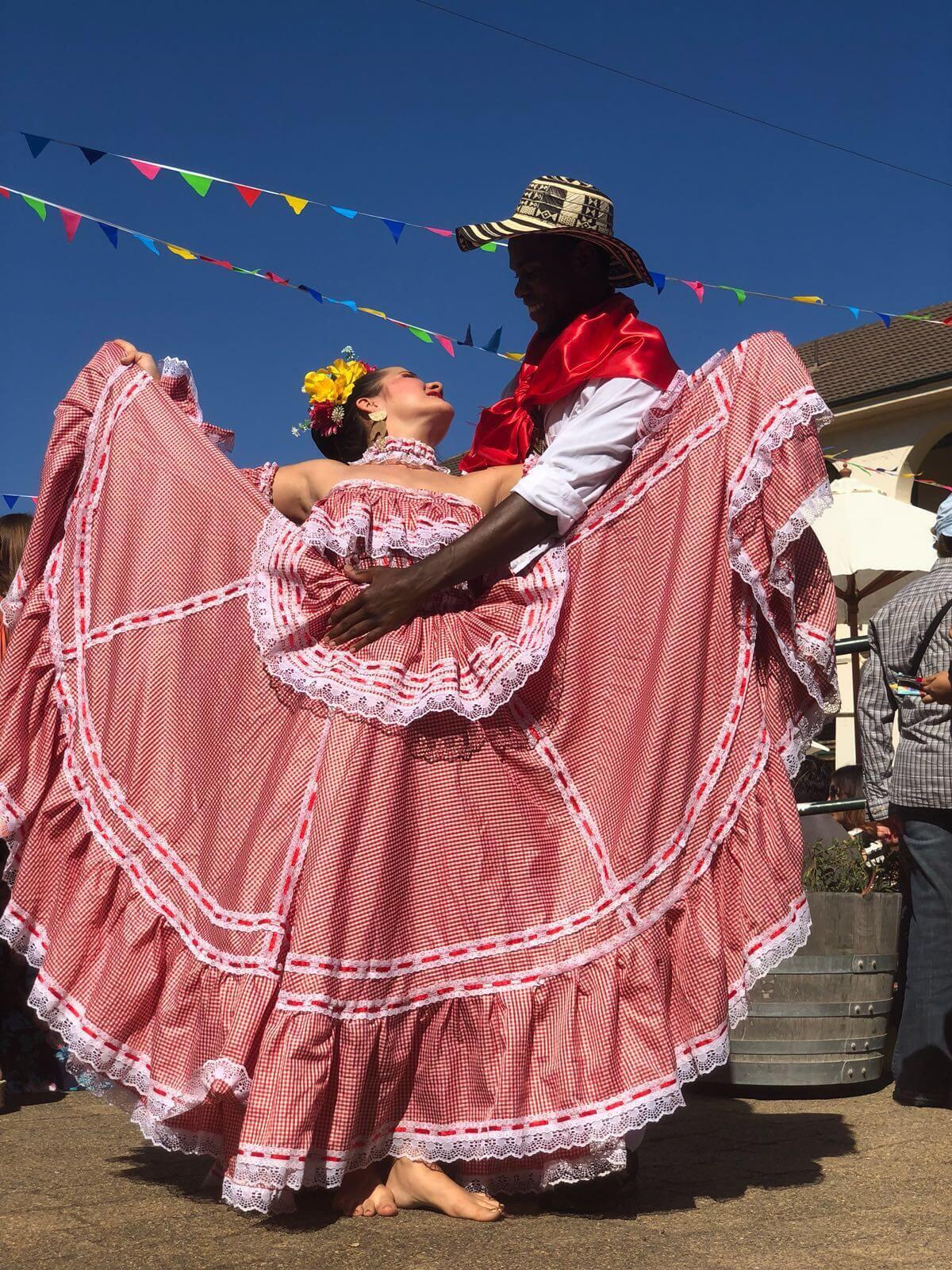 Una pareja baila alegremente con trajes coloridos. La mujer lleva un vestido tradicional rojo y blanco, y el hombre un sombrero y un pañuelo rojo. Por encima de ellos ondean banderas festivas.