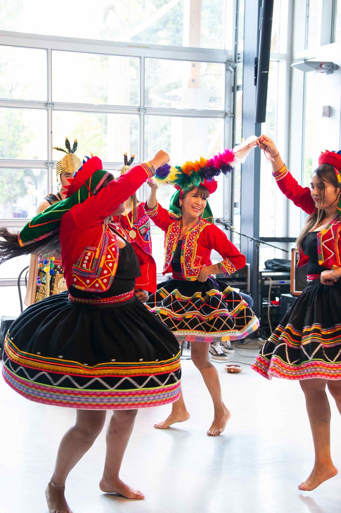 Cuatro bailarinas con vibrantes trajes tradicionales, con blusas rojas y faldas negras con estampados coloridos, bailan alegremente en el interior, transmitiendo una energía festiva.