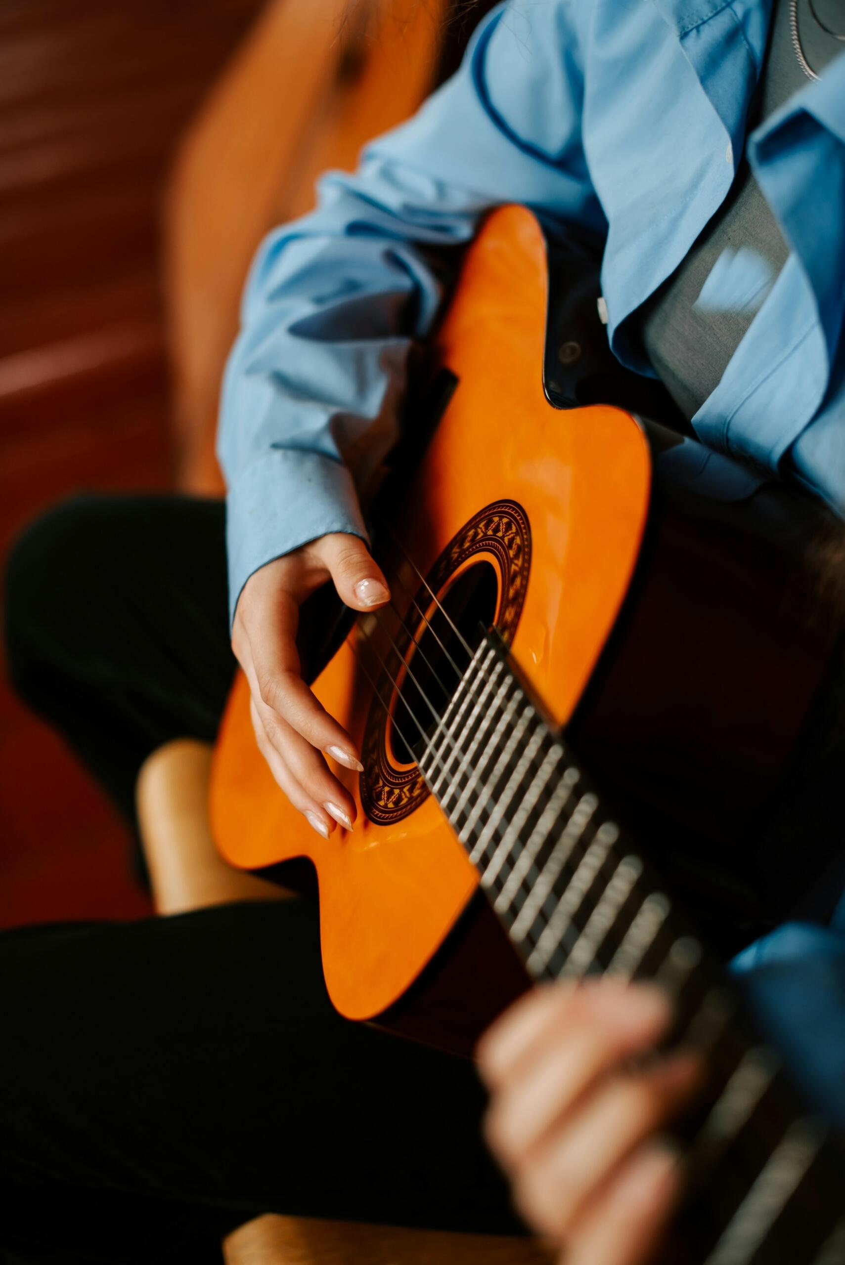 Persona con una camiseta azul tocando una guitarra acústica; el plano se centra en las manos que rasguean y tocan las cuerdas.