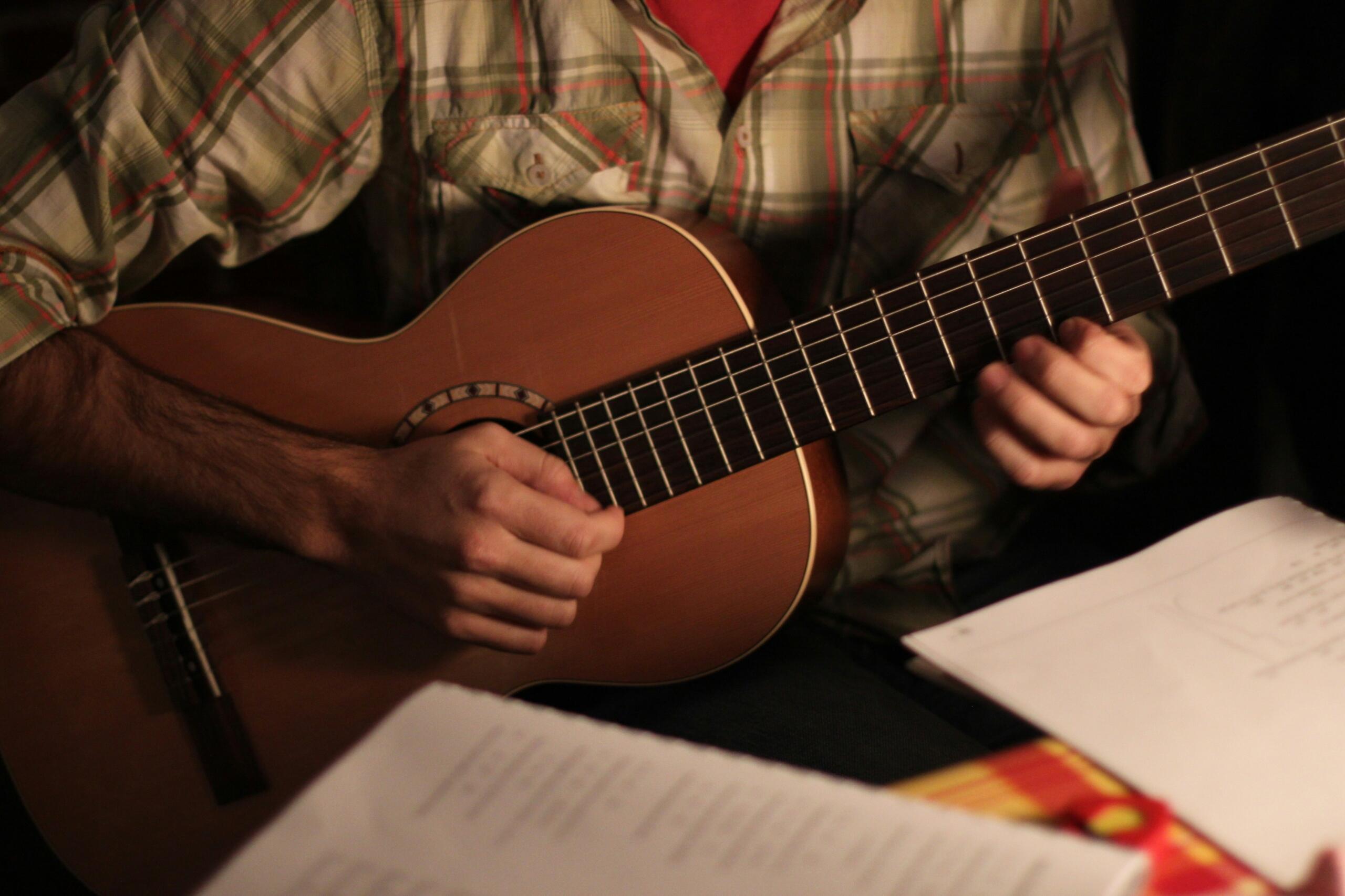 Una persona con una camisa a cuadros tocando la guitarra acústica, con una partitura en primer plano, bajo una luz tenue.
