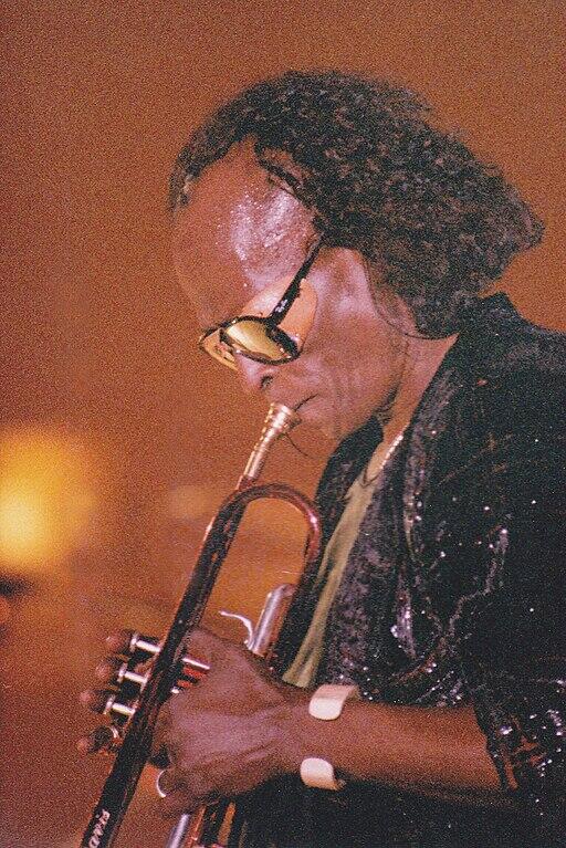Miles Davis toca con pasión la trompeta en el escenario, con gafas de sol y una chaqueta de lentejuelas. La cálida iluminación crea un ambiente íntimo y evocador.