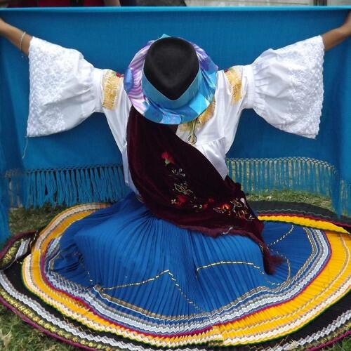 Una persona con un vibrante atuendo tradicional posa con elegancia. Lleva un sombrero de ala ancha, una falda colorida de varias capas, un chal bordado y sostiene una tela azul brillante con los brazos abiertos, irradiando orgullo cultural.
