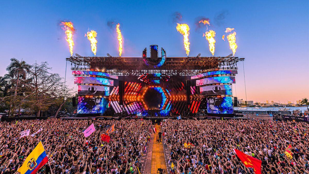 Un animado festival de música con un gran escenario con luces de colores y pirotecnia, y un público entusiasta ondeando banderas bajo un cielo despejado.