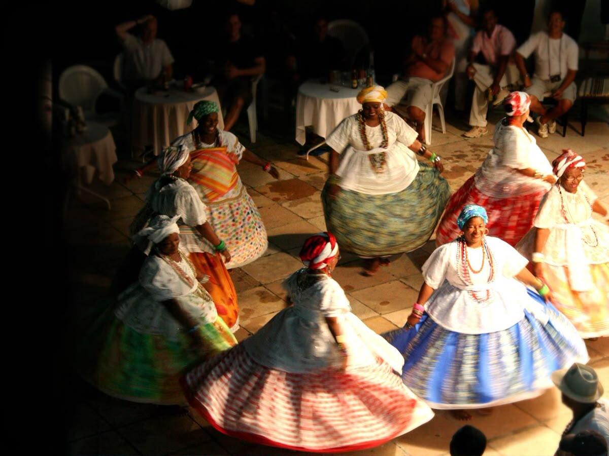 Un grupo de bailarinas con vestidos coloridos y fluidos y pañuelos en la cabeza bailan en círculo. Están sobre un suelo de baldosas, observadas por espectadores sentados.