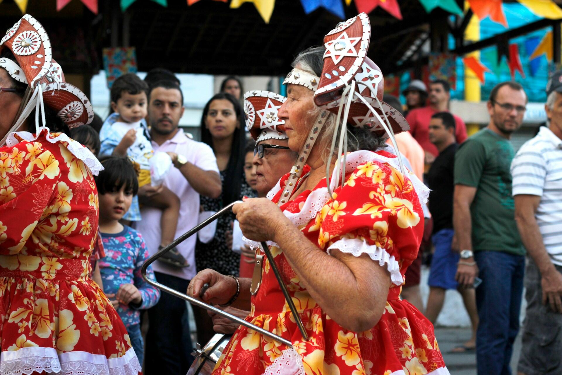 Mujeres mayores con vestidos florales de colores vivos, rojos y amarillos, tocan triángulos en un animado festival callejero, con un público diverso y participativo al fondo.