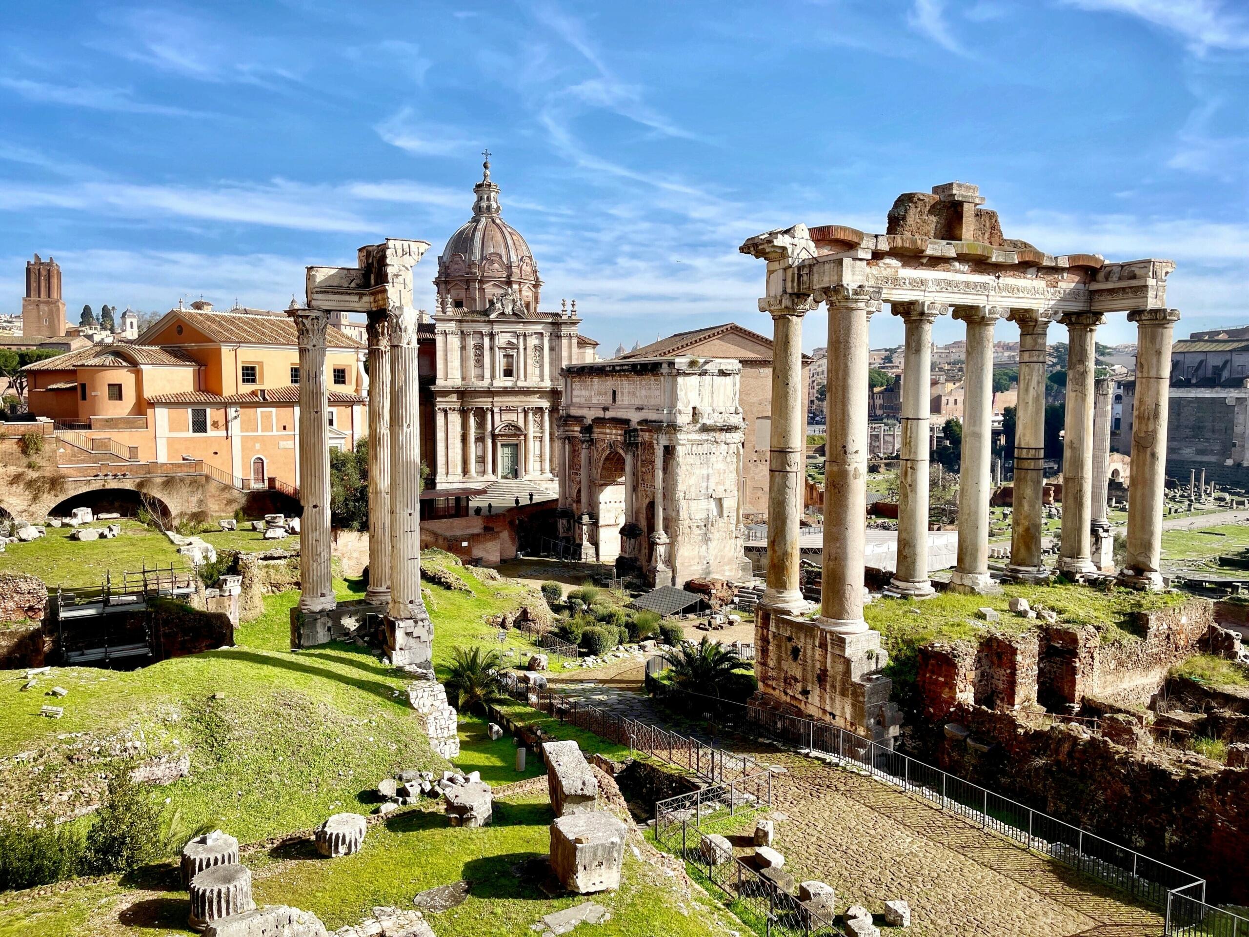 Las ruinas del antiguo Foro Romano, con sus imponentes columnas y las ornamentadas cúpulas de las iglesias, bajo un cielo azul brillante.