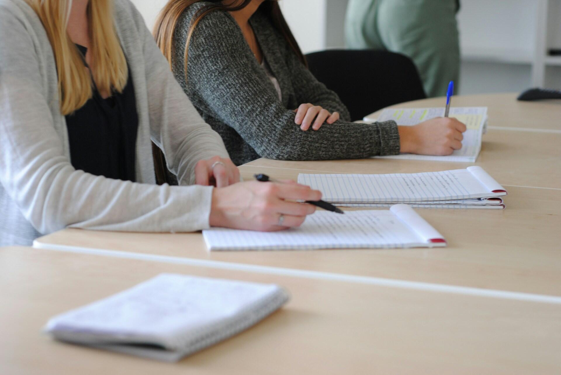Los alumnos toman apuntes en cuadernos abiertos sobre las mesas del aula, lo que crea un ambiente de concentración y colaboración durante el estudio o la clase.
