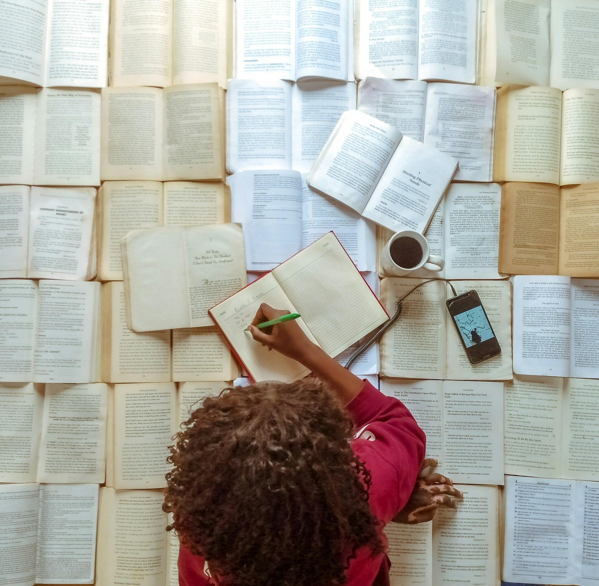 Vista aérea de una persona con una camiseta roja que escribe en un cuaderno abierto rodeada de libros, café y un teléfono, creando una escena de estudio en la que se nota la concentración.