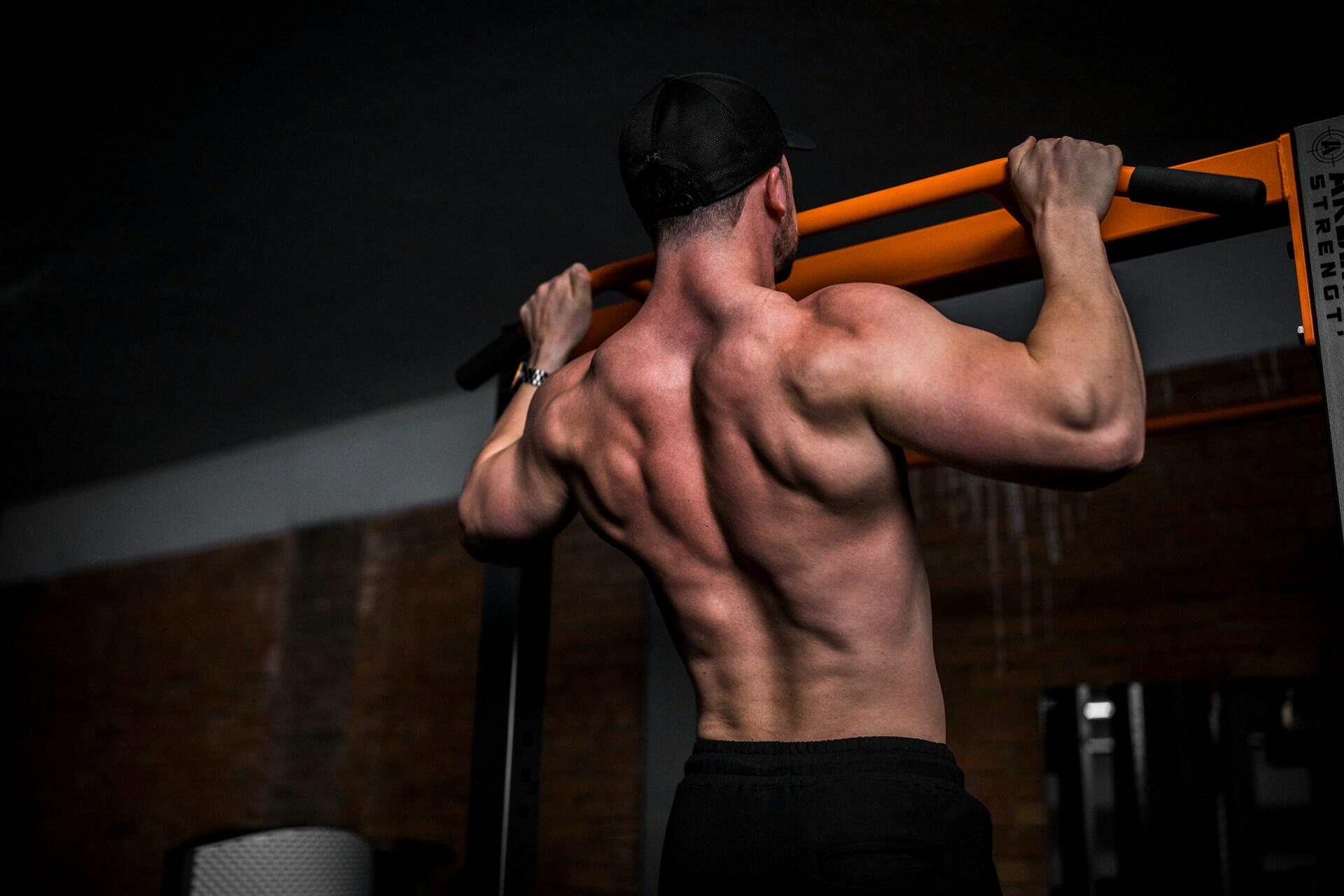 Hombre musculoso haciendo una dominada en una barra naranja, con gorra negra y pantalones cortos, en un gimnasio. Se destaca su espalda robusta, lo que transmite fuerza.
