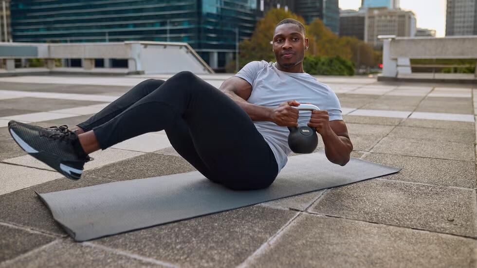 Un hombre haciendo ejercicio al aire libre en una azotea, realizando un «Russian twist» con una pesa rusa. Está sobre una esterilla de yoga, rodeado de modernos edificios de la ciudad.