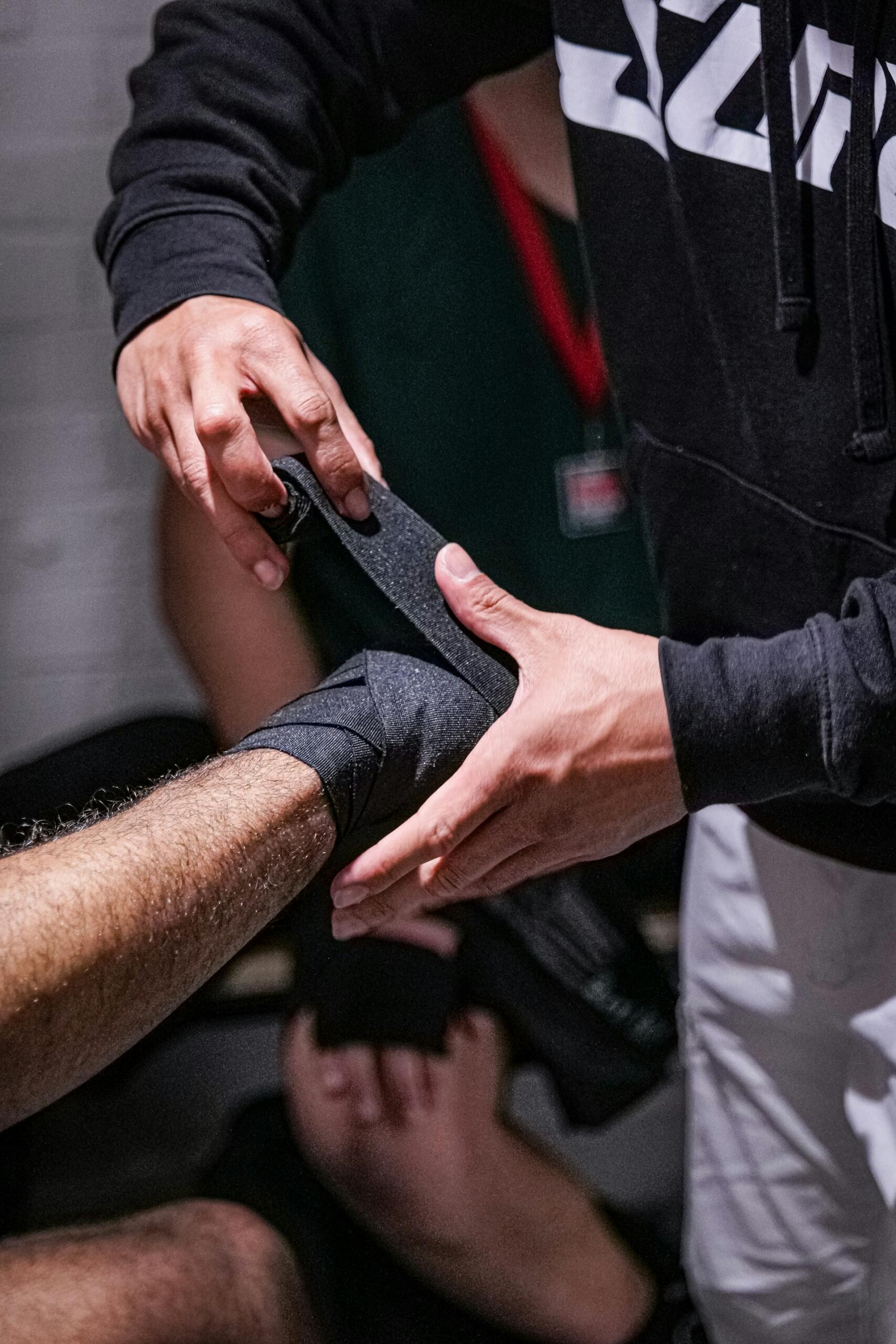 Una persona que lleva una sudadera con capucha negra envuelve con destreza una venda deportiva negra alrededor del tobillo de un deportista, lo que sugiere una preparación y un cuidado minuciosos.
