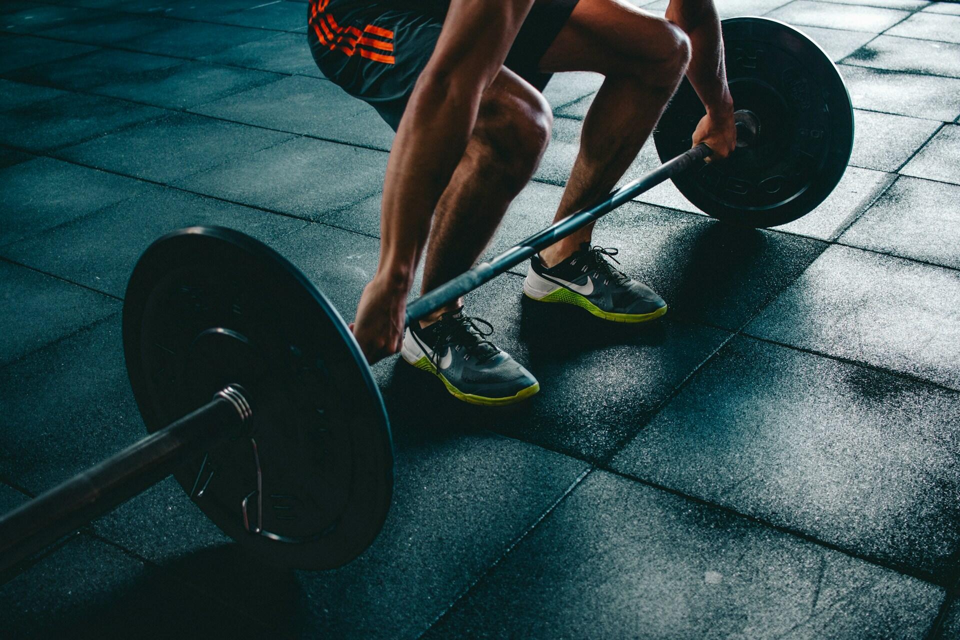 Una persona está agachada, sujetando una barra en el suelo oscuro de un gimnasio; lleva una camiseta gris y unos pantalones cortos con detalles en verde neón. La escena transmite concentración y fuerza.
