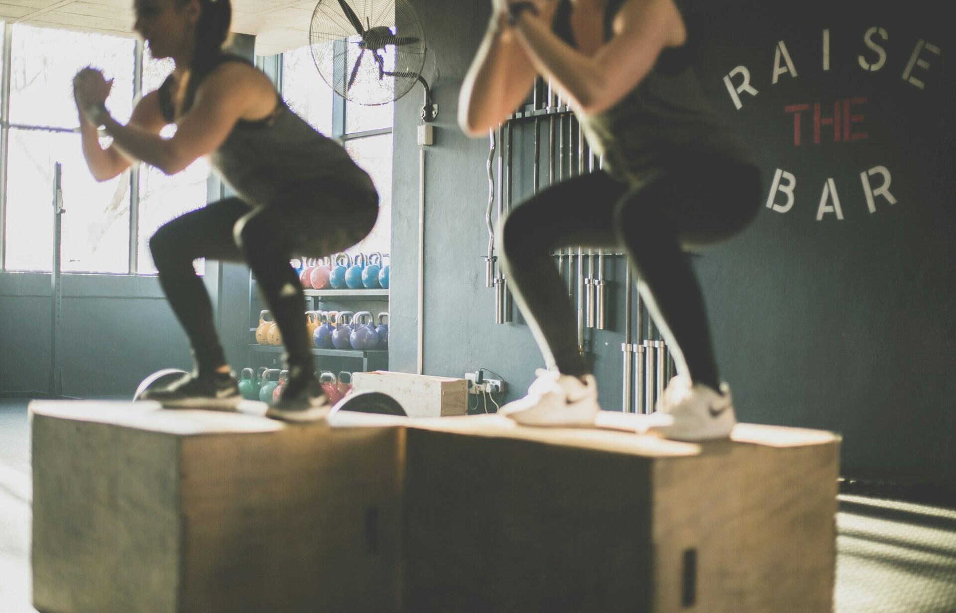 Dos personas están haciendo sentadillas sobre cajas de madera en un gimnasio. La sala está bien iluminada gracias a unos grandes ventanales, y al fondo se ven unas pesas rusas de colores.