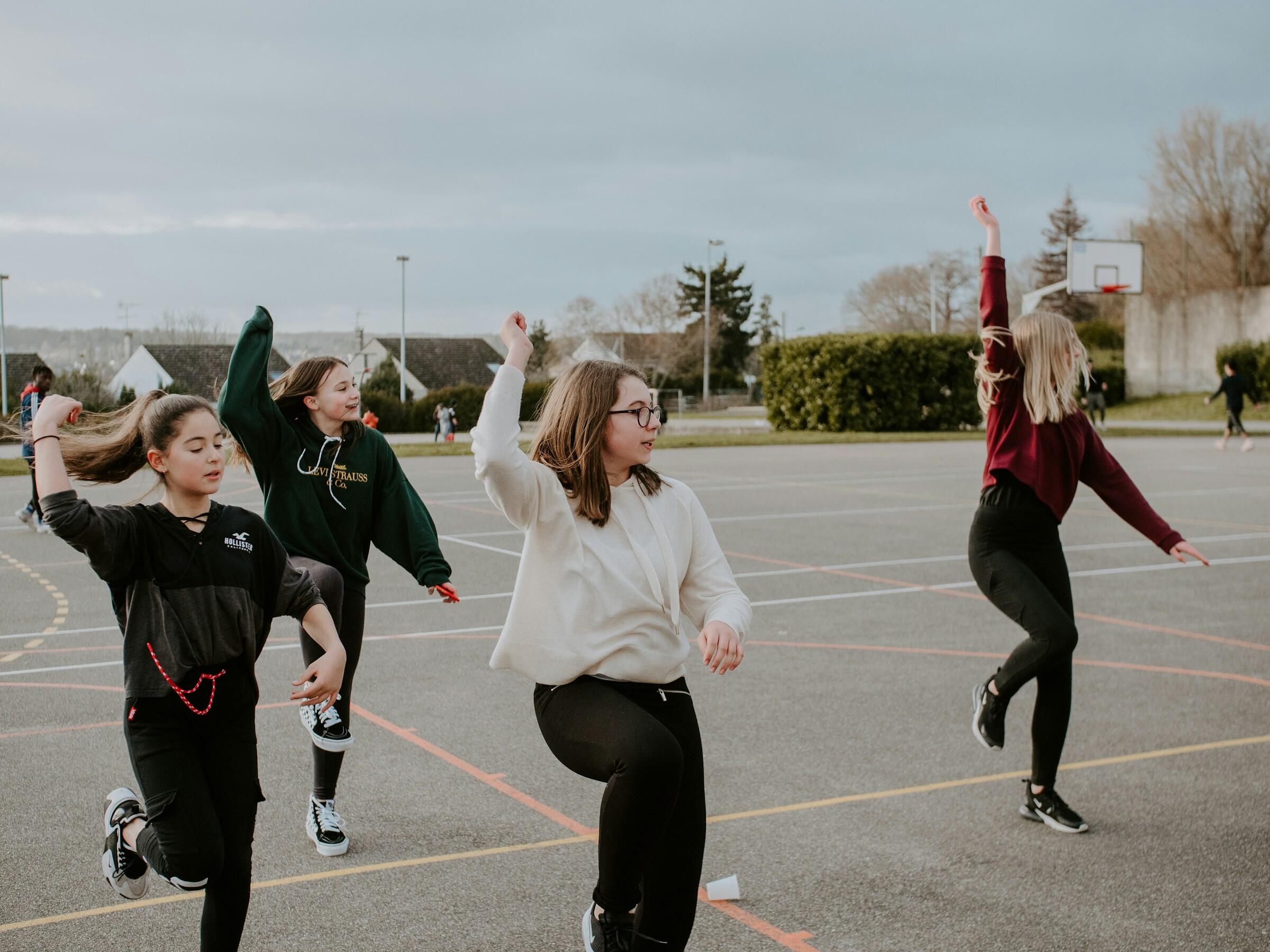 Cuatro chicas bailan con energía en una cancha de baloncesto al aire libre, bajo un cielo nublado.