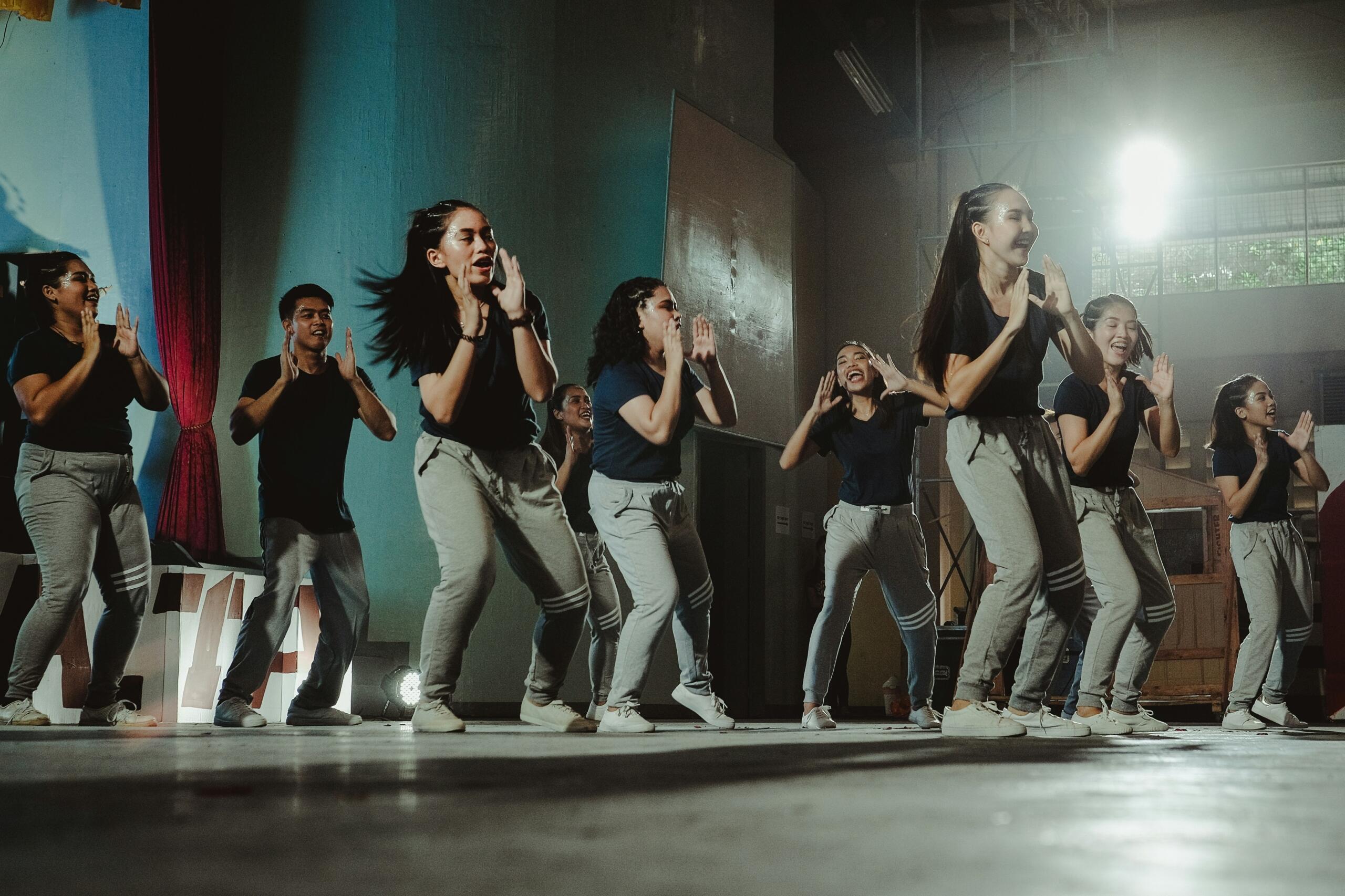 Un grupo de jóvenes con ropa a juego que bailan con energía en el escenario, bajo las luces brillantes de una sala cubierta.