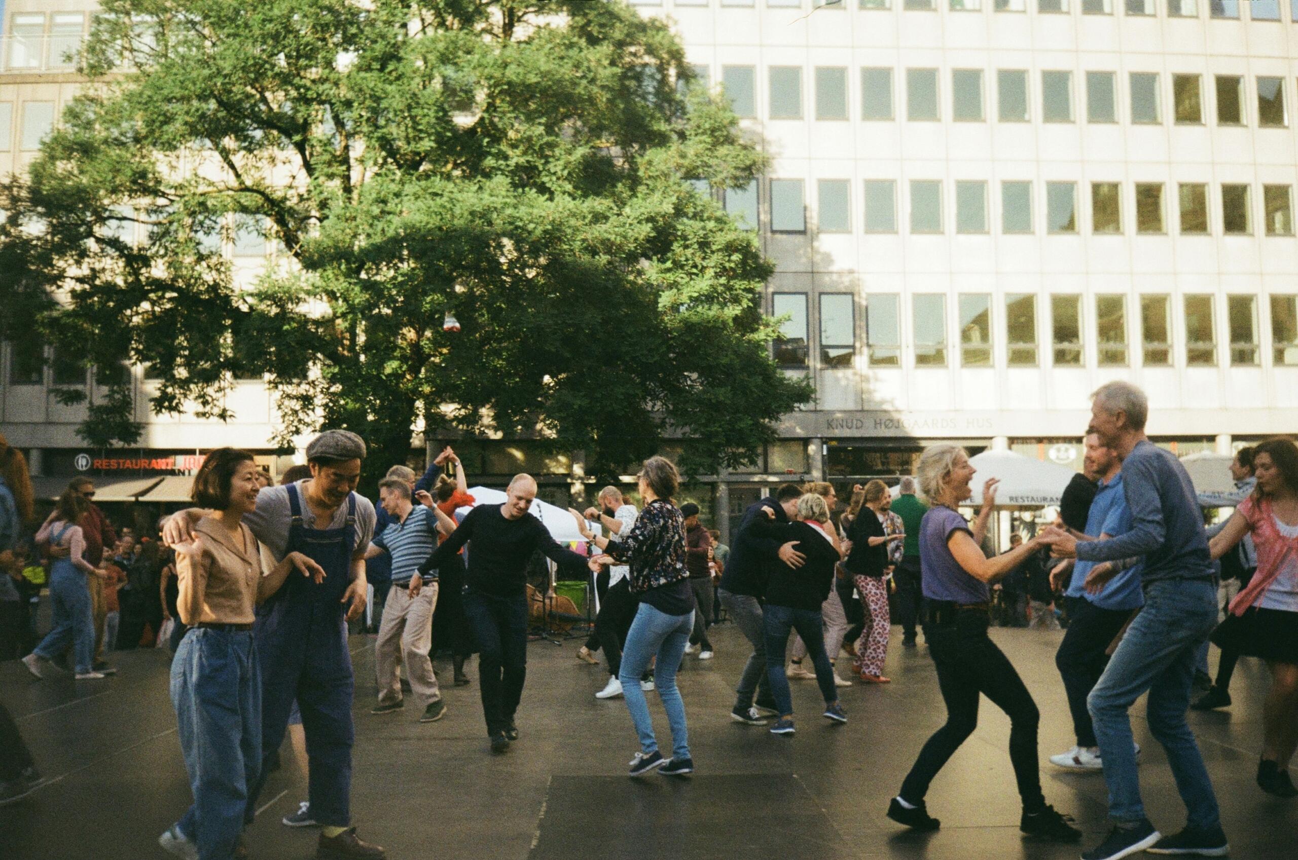 Gente bailando alegremente al aire libre en una plaza de la ciudad a plena luz del día, rodeada de árboles y de un edificio de oficinas de varias plantas.