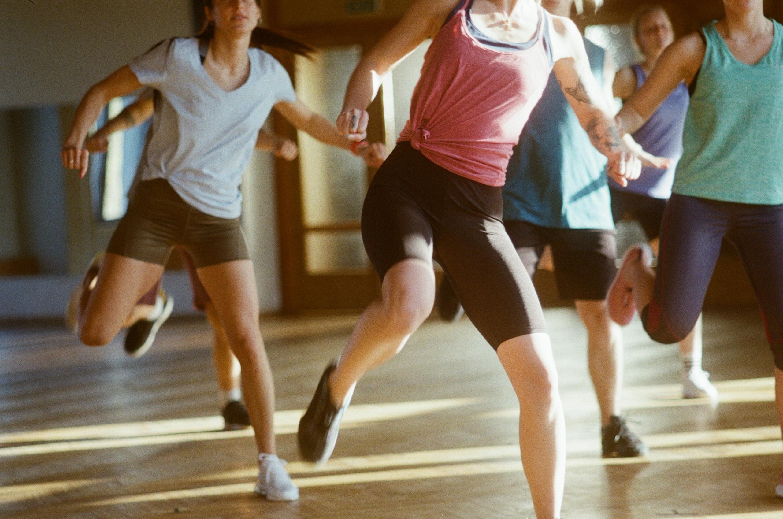 Un grupo de personas con ropa deportiva haciendo ejercicio en el interior, realizando un movimiento dinámico de elevación de piernas en una sala iluminada por la luz del sol.