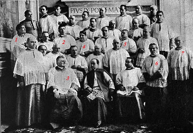 Un grupo de clérigos, ataviados con vestimentas tradicionales, posa solemnemente en el interior de una gran sala. El fondo presenta detalles arquitectónicos y la inscripción «PIUS IX PONT MAX». El ambiente es formal e histórico.