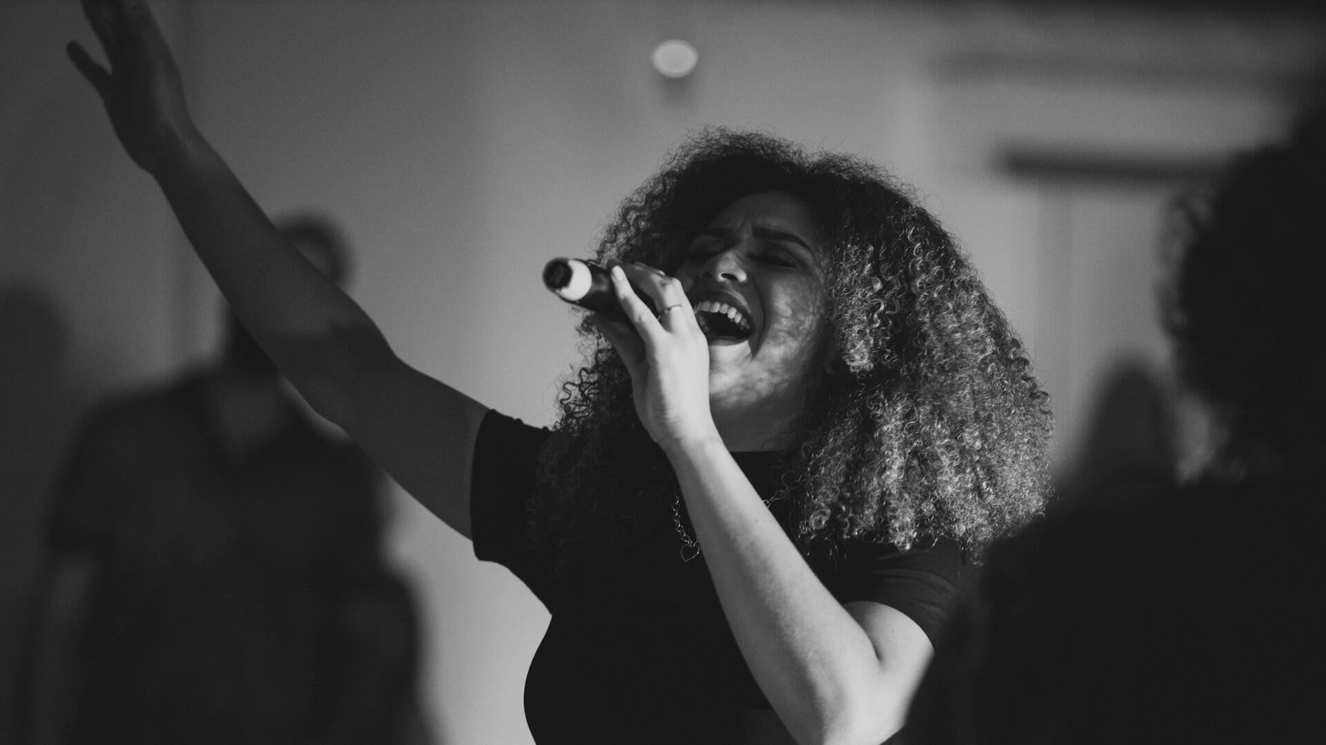 Una mujer canta con pasión ante un micrófono, con la mano levantada y los ojos cerrados. La imagen en blanco y negro capta un momento musical intenso sobre el escenario.