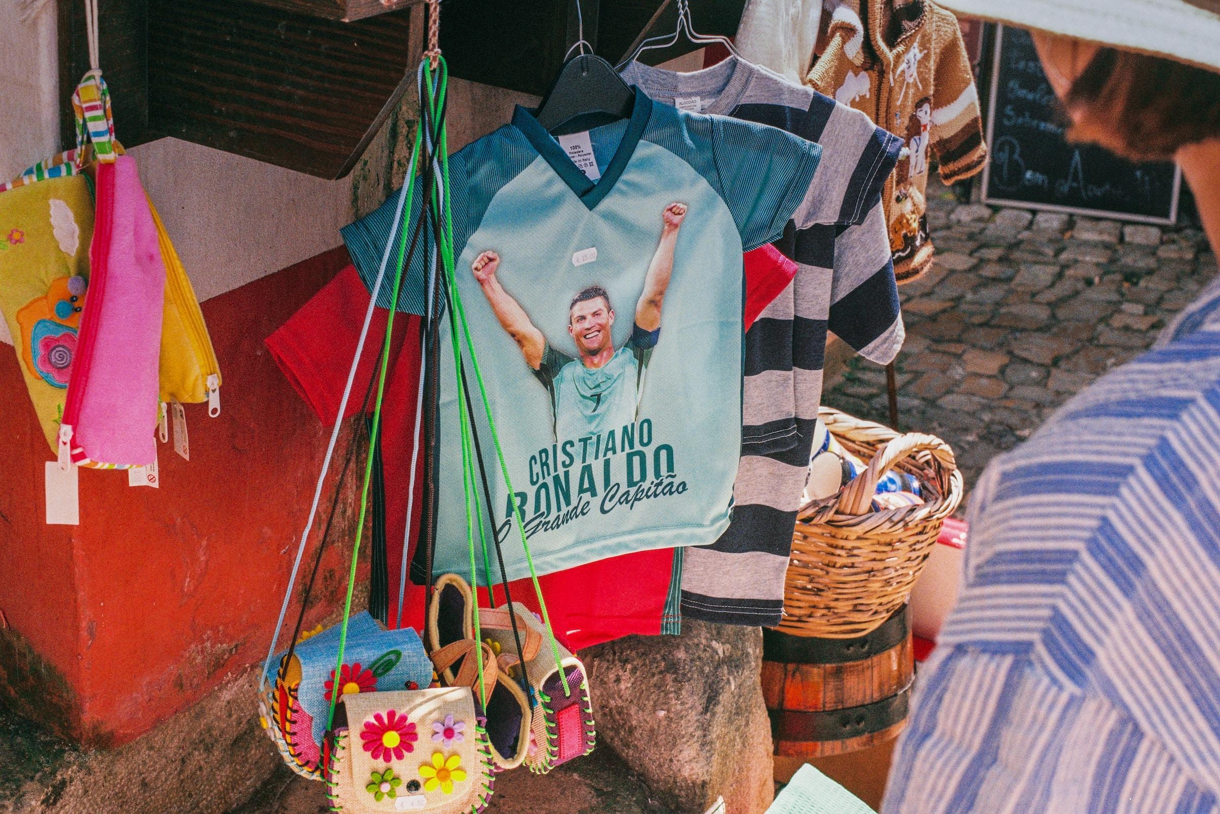 Un puesto del mercado exhibe una camiseta azul con la imagen de Cristiano Ronaldo con los brazos en alto. A su alrededor hay bolsos de colores y camisetas a rayas, lo que le da un aire vibrante y alegre.