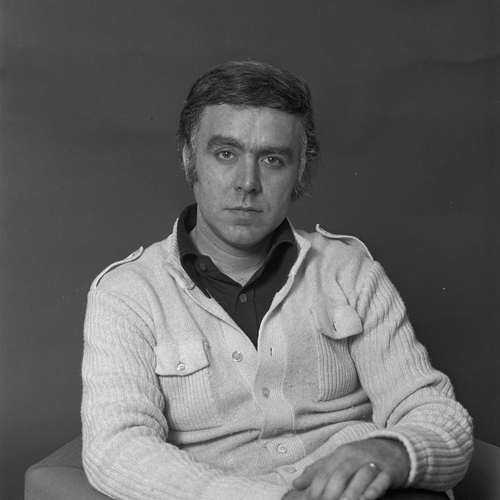 Foto en blanco y negro de Carlos do Carmo sentado con un jersey claro sobre una camisa oscura, con expresión neutra, sobre un fondo oscuro sencillo.