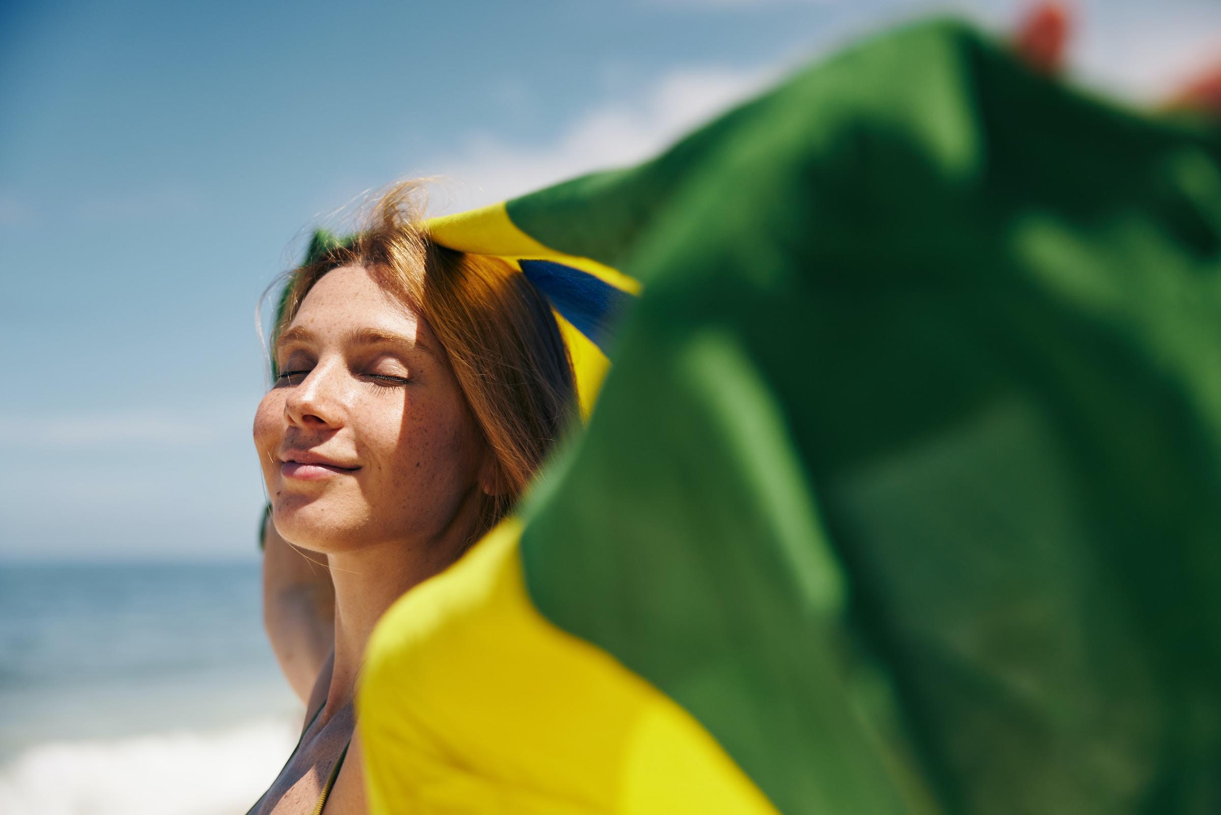 Una mujer sonríe con los ojos cerrados y sostiene una bandera de Brasil con una playa soleada y luminosa como telón de fondo. El ambiente es sereno y alegre.
