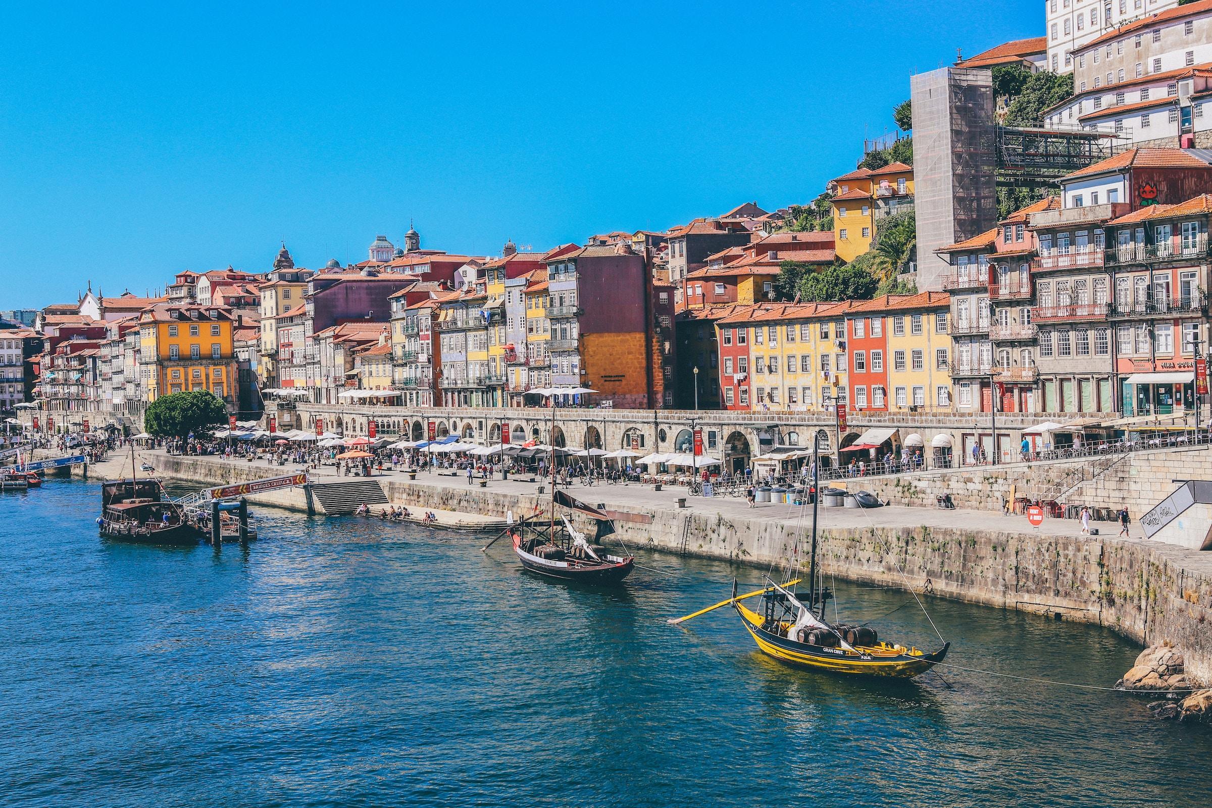 Edificios coloridos se alinean a lo largo de la ribera del río en Oporto, Portugal, con barcos tradicionales balanceándose en las aguas azules bajo un cielo despejado.
