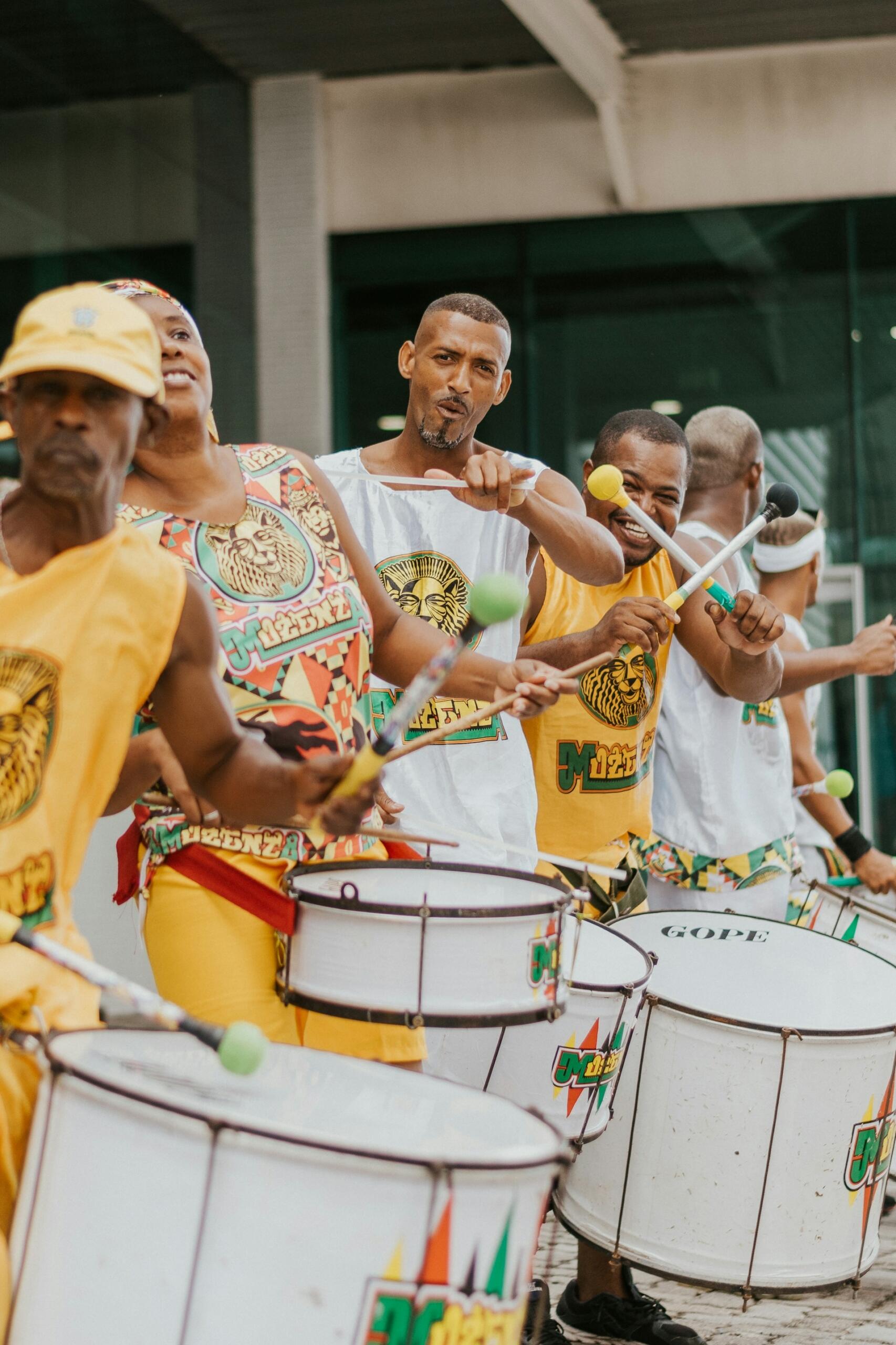 Un grupo de músicos sonrientes con trajes coloridos y sombreros amarillos tocan grandes tambores blancos con baquetas verdes y amarillas, irradiando alegría y ritmo.