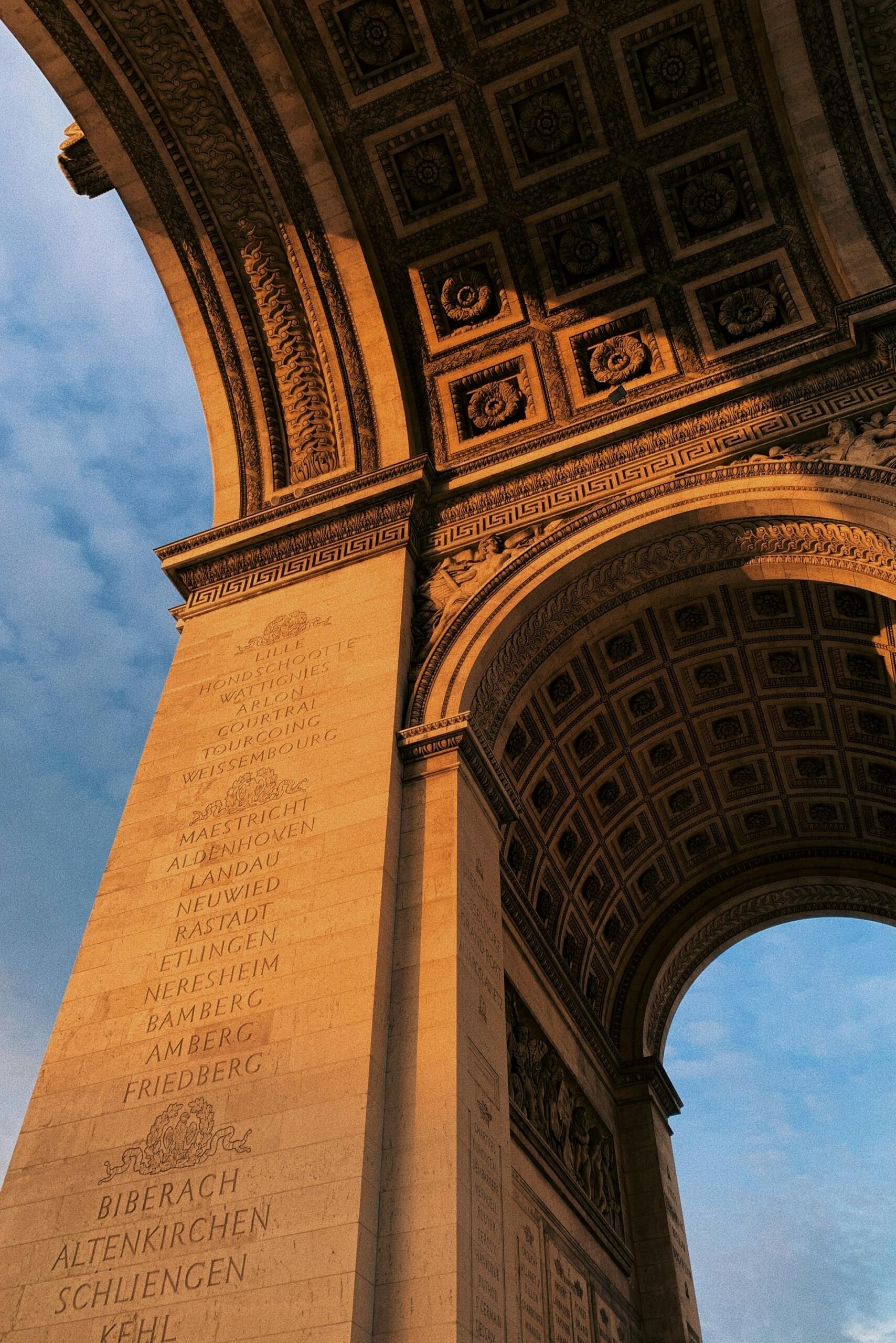 Vista del Arco del Triunfo desde abajo, con sus elaboradas tallas e inscripciones iluminadas por la cálida luz del atardecer. El cielo está parcialmente nublado y azul.