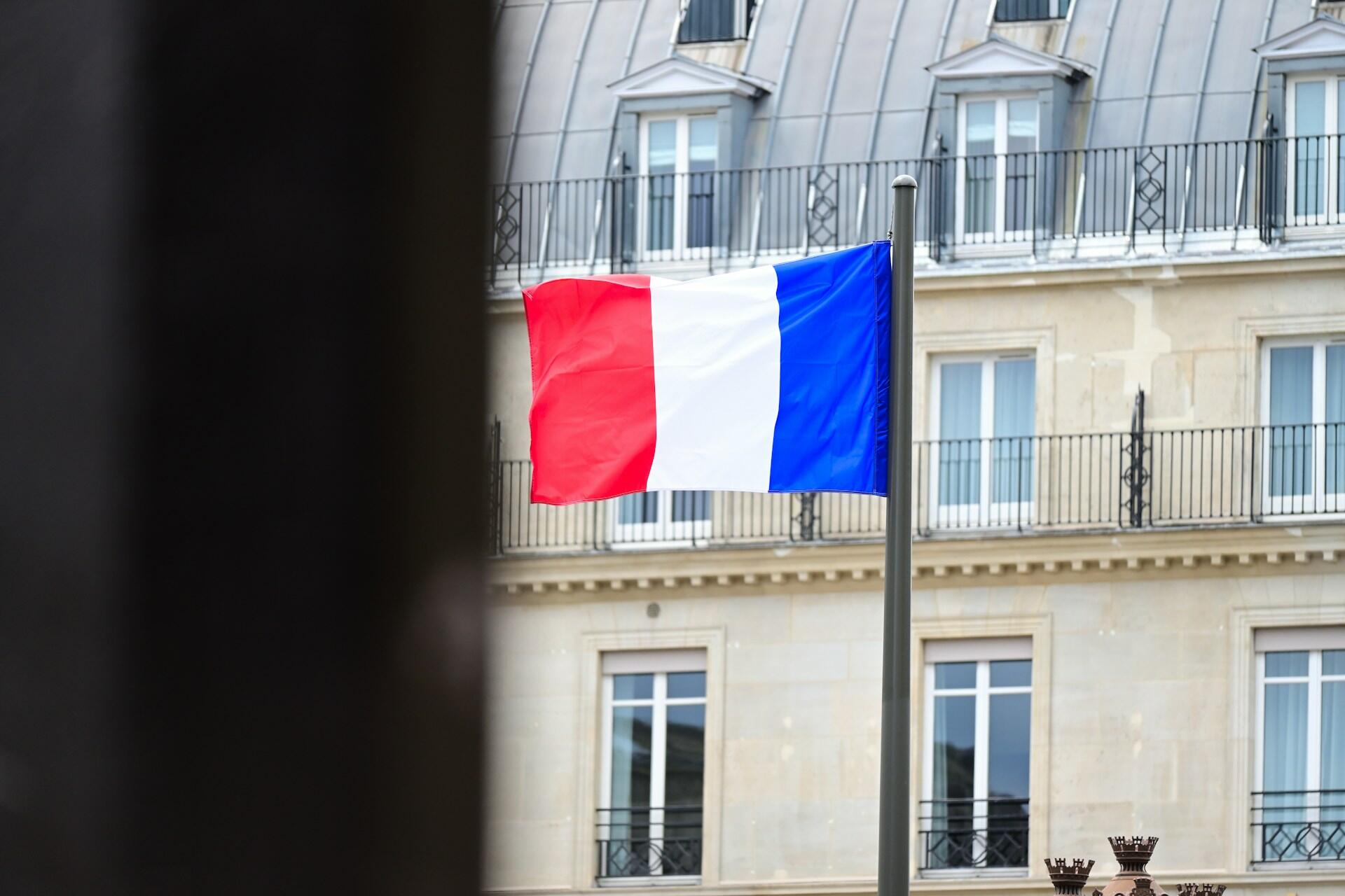 La bandera francesa ondea en un mástil frente a un elegante edificio con tejado abuhardillado. La escena transmite una sensación de orgullo nacional y sofisticación.