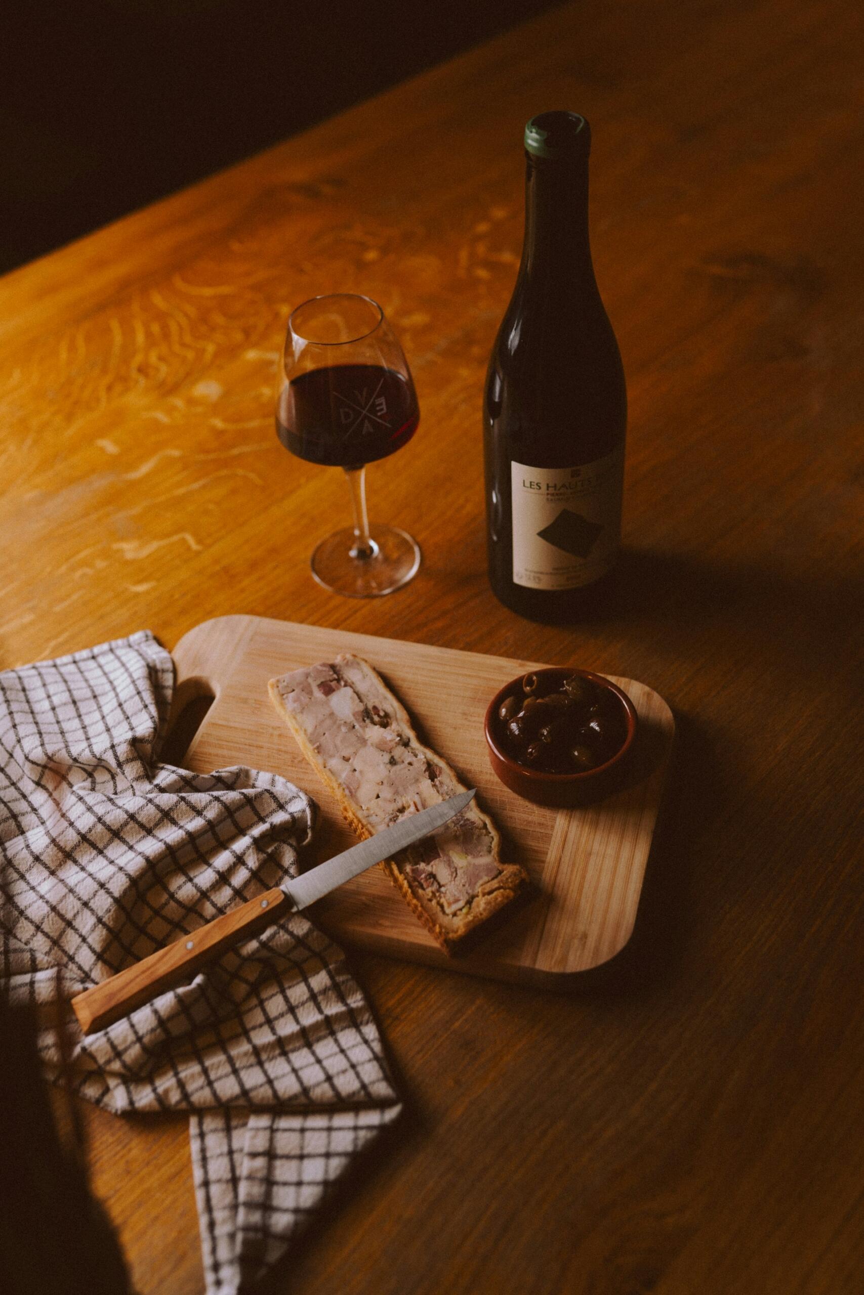Una mesa rústica de madera con una botella de vino, una copa de vino tinto, una tabla de cortar con paté, aceitunas, un cuchillo y un mantel a cuadros. Ambiente evocador.