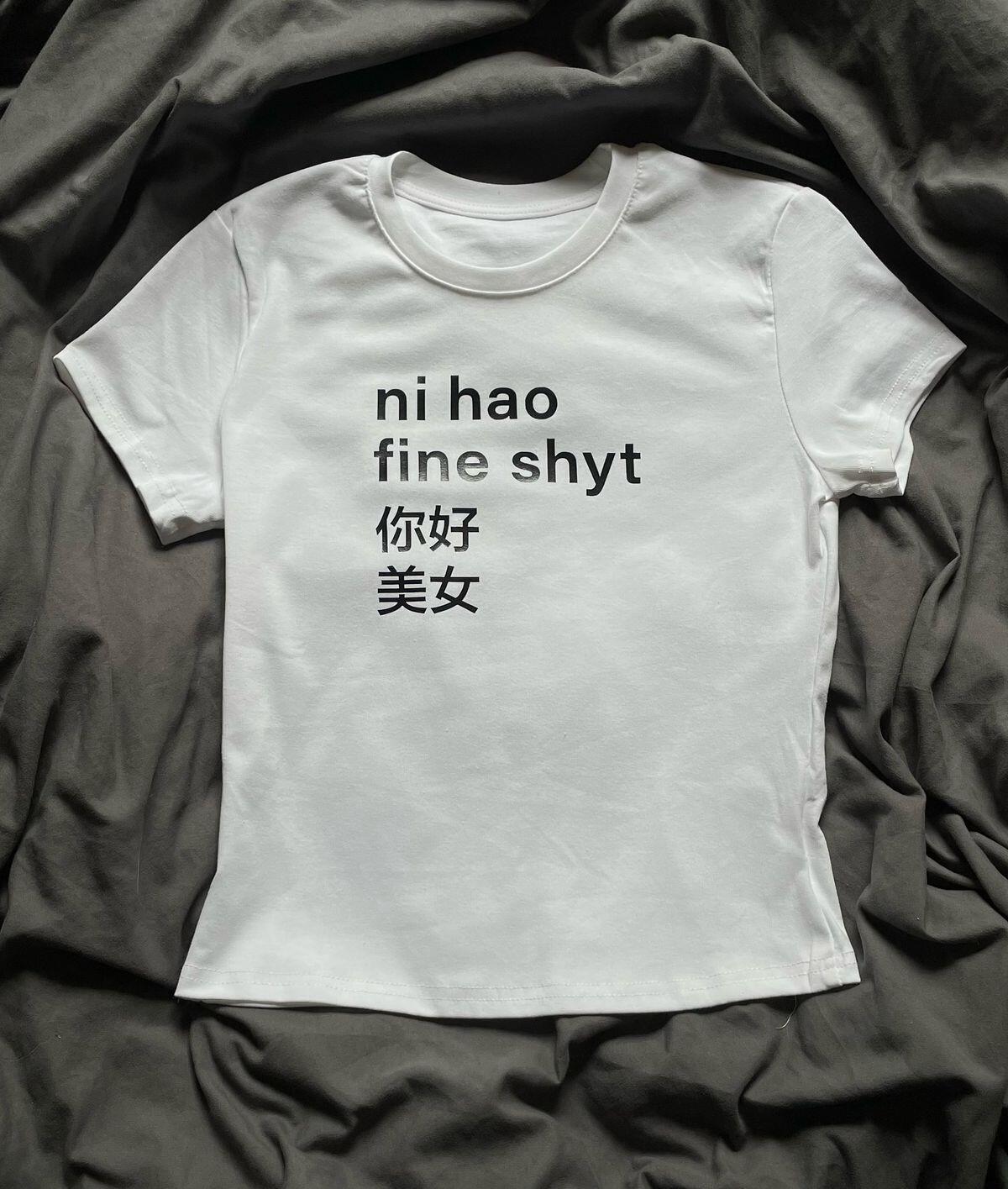 Camiseta blanca sobre una tela gris arrugada, con un llamativo texto en negro: «ni hao fine shyt», seguido de unos caracteres chinos. Tono informal y desenfadado.