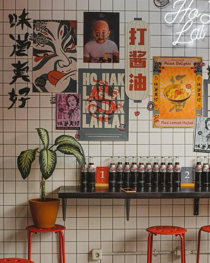 Un acogedor restaurante con paredes de azulejos blancos decoradas con llamativos pósters asiáticos y un letrero de neón. Unos taburetes rojos se alinean junto a una estantería negra en la que se exponen botellas de condimentos, junto a una planta en maceta, creando un ambiente animado y acogedor.